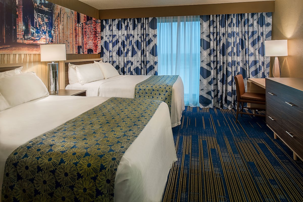 Habitación de hotel con dos camas, un escritorio, una silla y un ventanal con cortinas.
