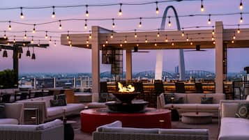 An outdoor seating area with wicker furniture, a central fire feature, and string lights against a cityscape backdrop.