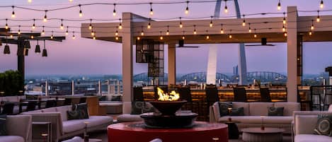 An outdoor seating area with wicker furniture, a central fire feature, and string lights against a cityscape backdrop.