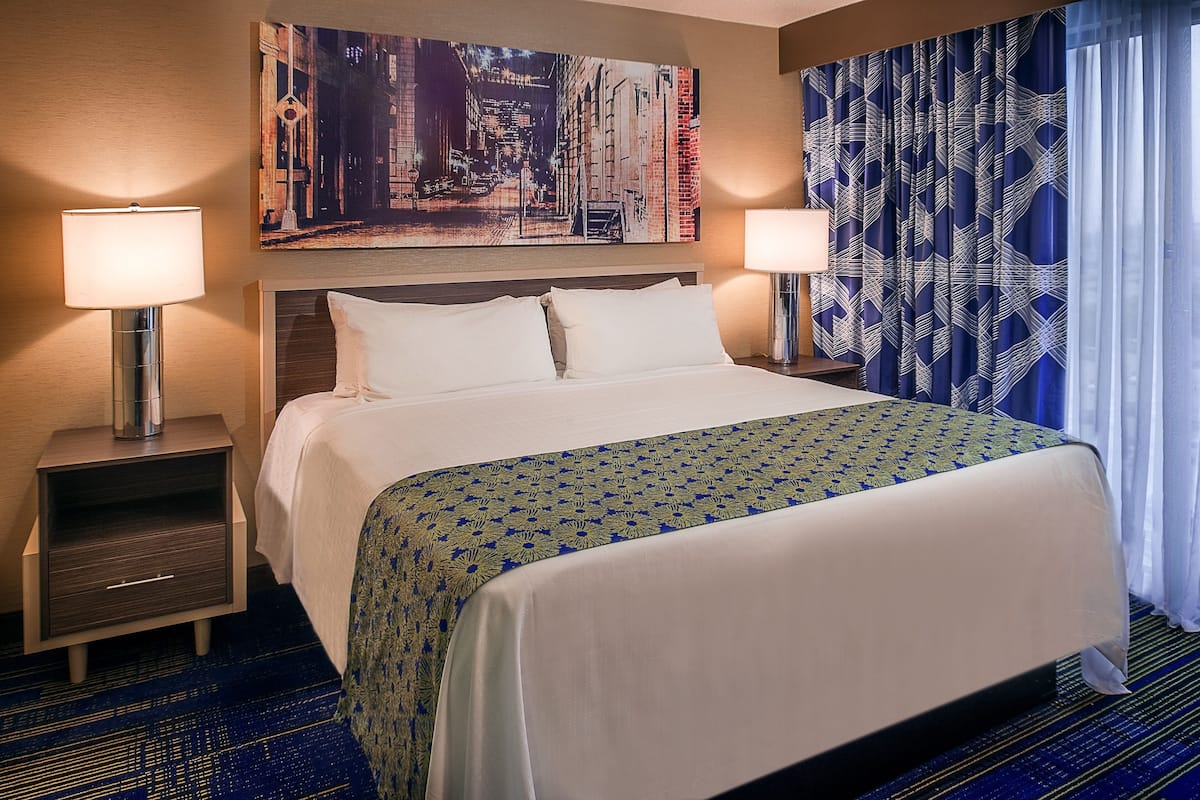 Habitación de hotel con una cama grande, dos mesitas de noche con lámparas, un cabecero, un cuadro de un paisaje urbano, cortinas estampadas y piso alfombrado.