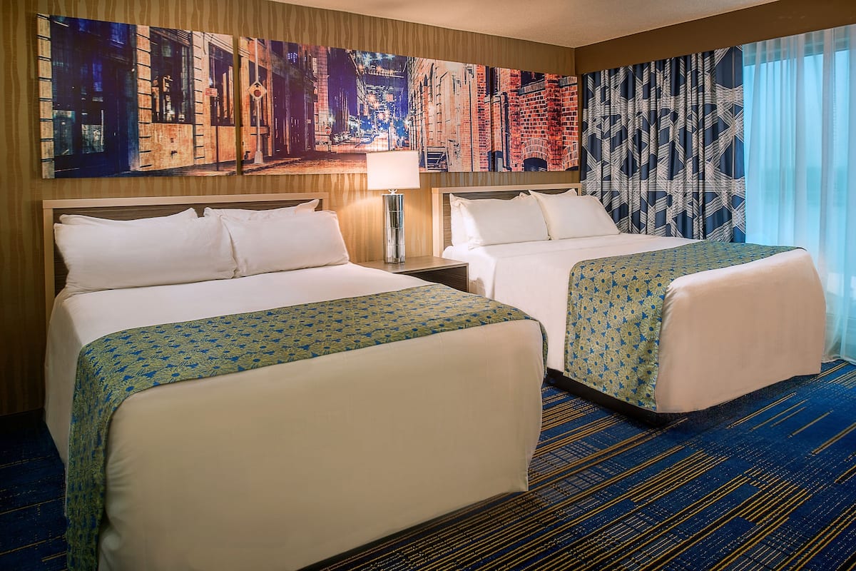 Una habitación de hotel con dos camas, un cuadro de gran formato con un paisaje urbano en la pared, una alfombra estampada y una ventana con cortinas.