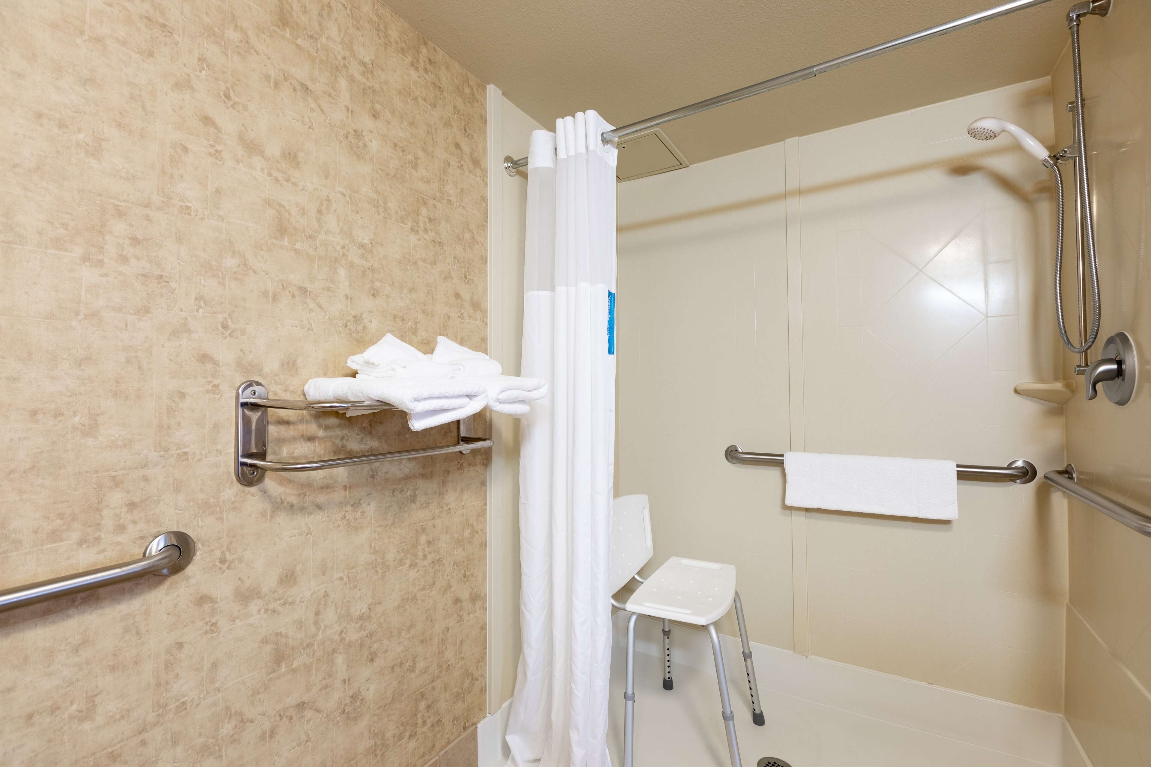 Une salle de bain équipée d’une douche, de barres d’appui, d’une chaise blanche et d’un porte-serviettes.