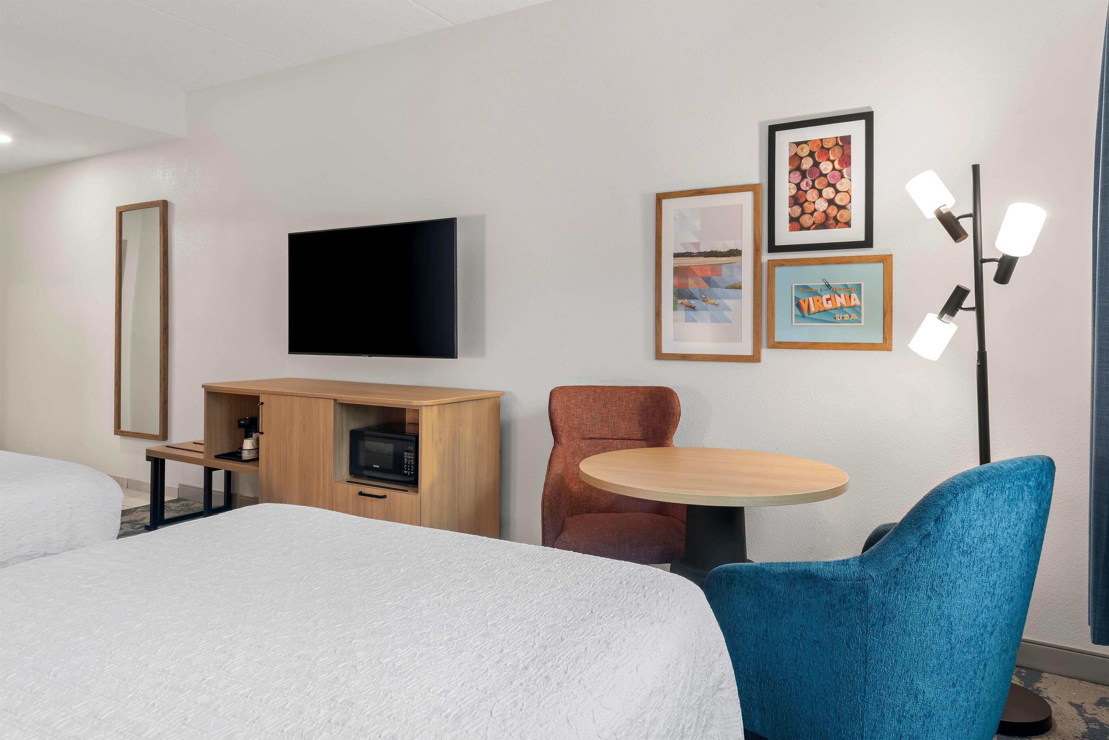 Habitación de hotel con cama, televisor, aparador de madera, mesa redonda y una silla azul.