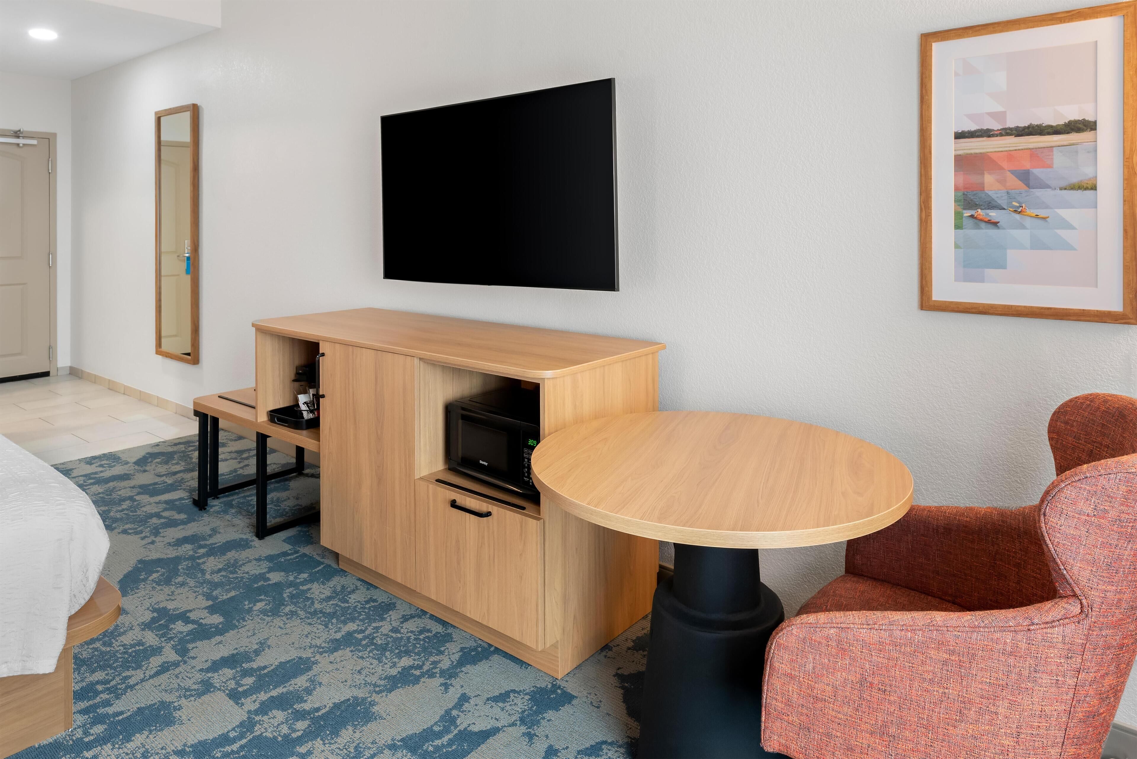 Habitación de hotel con un mueble de madera, un televisor de pantalla plana, una mesa redonda de madera, un sillón tapizado rojo y un cuadro en la pared.