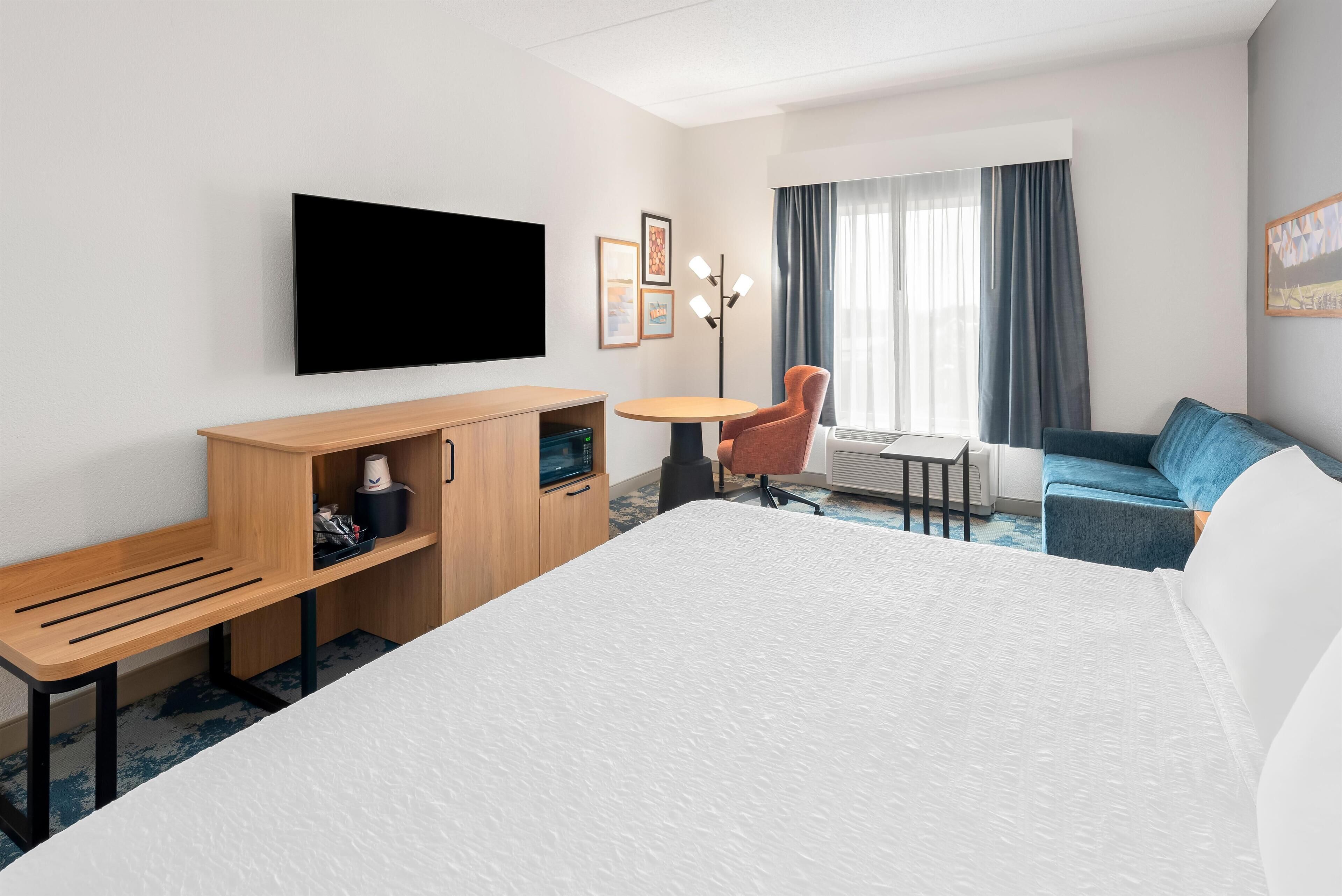 Une chambre d’hôtel avec un grand lit, un bureau en bois, une chaise, une petite table, un téléviseur fixé au mur et une fenêtre avec des rideaux.