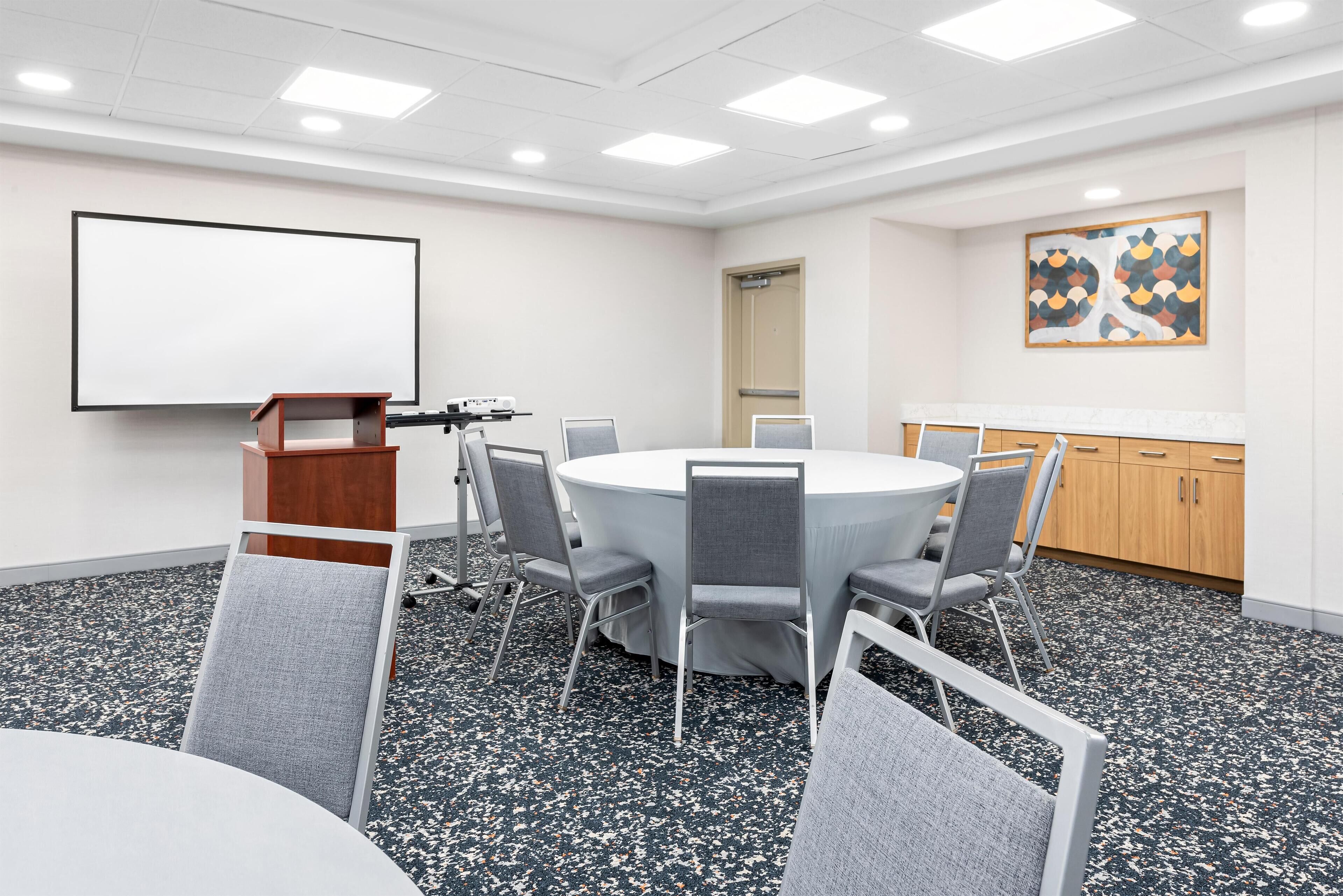 A conference room with a round table, grey chairs, a podium, a projector screen, and a painting on the wall.