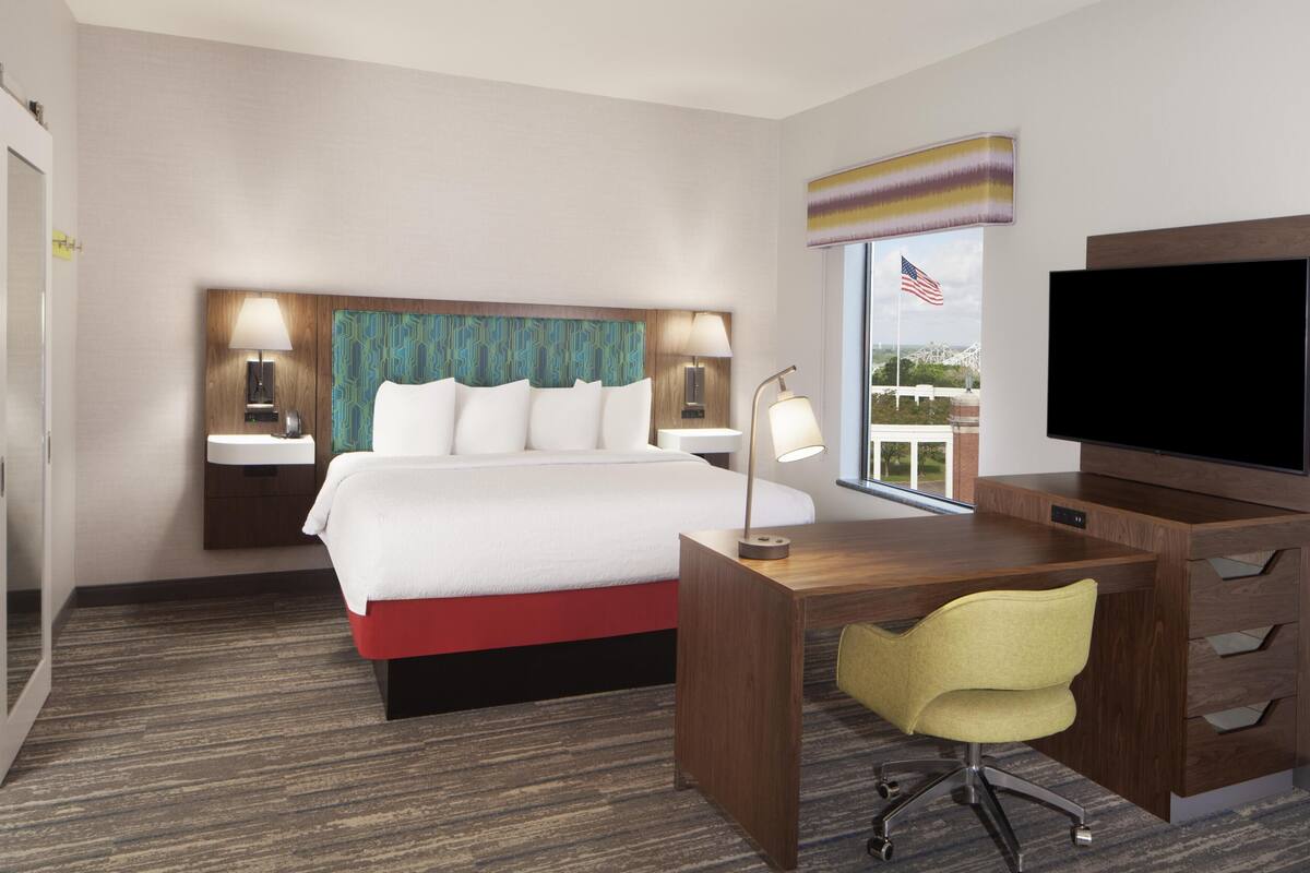 A hotel room with a bed, desk, chair, TV, and a window with an American flag.