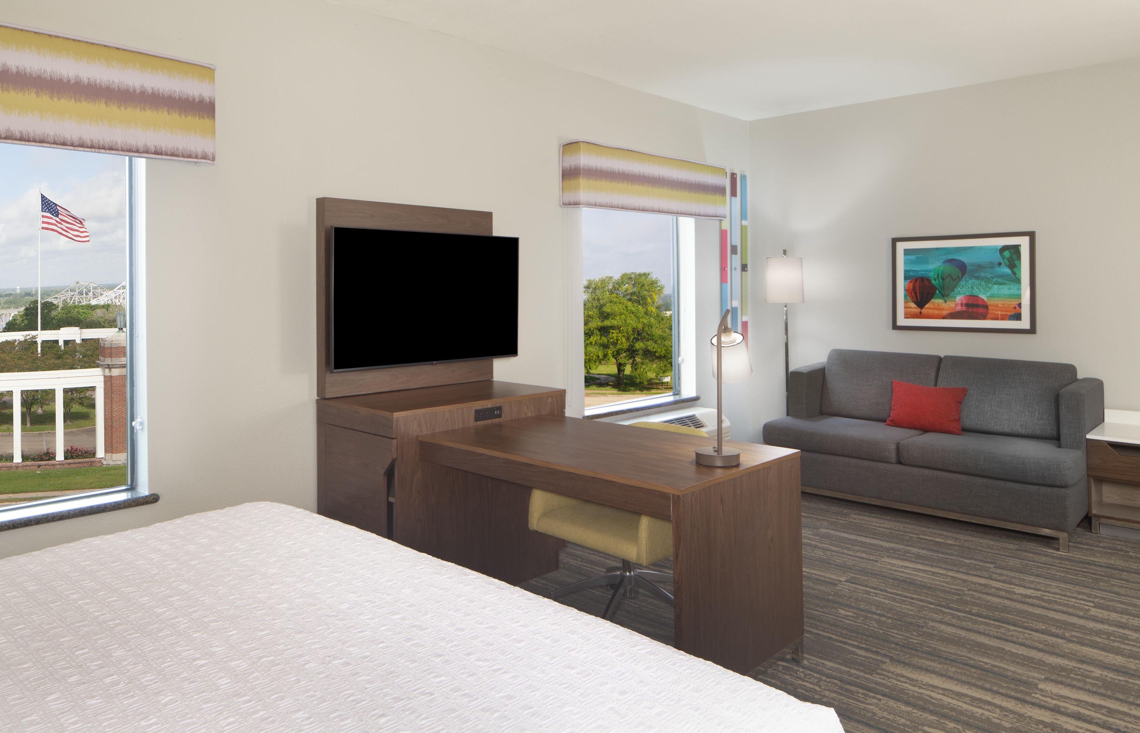 A hotel room with a bed, a desk, a television, a sofa, and a window with an American flag.