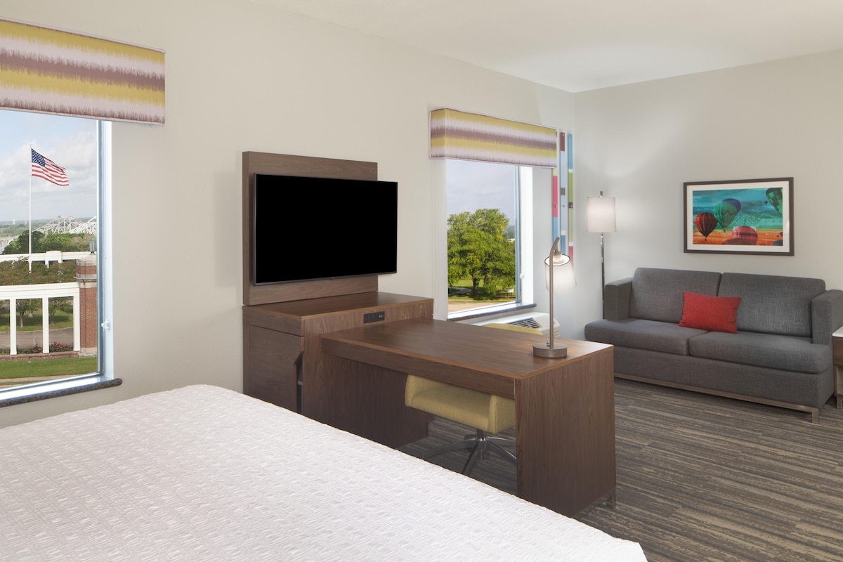 A hotel room with a bed, a desk, a television, a sofa, and a window with an American flag.