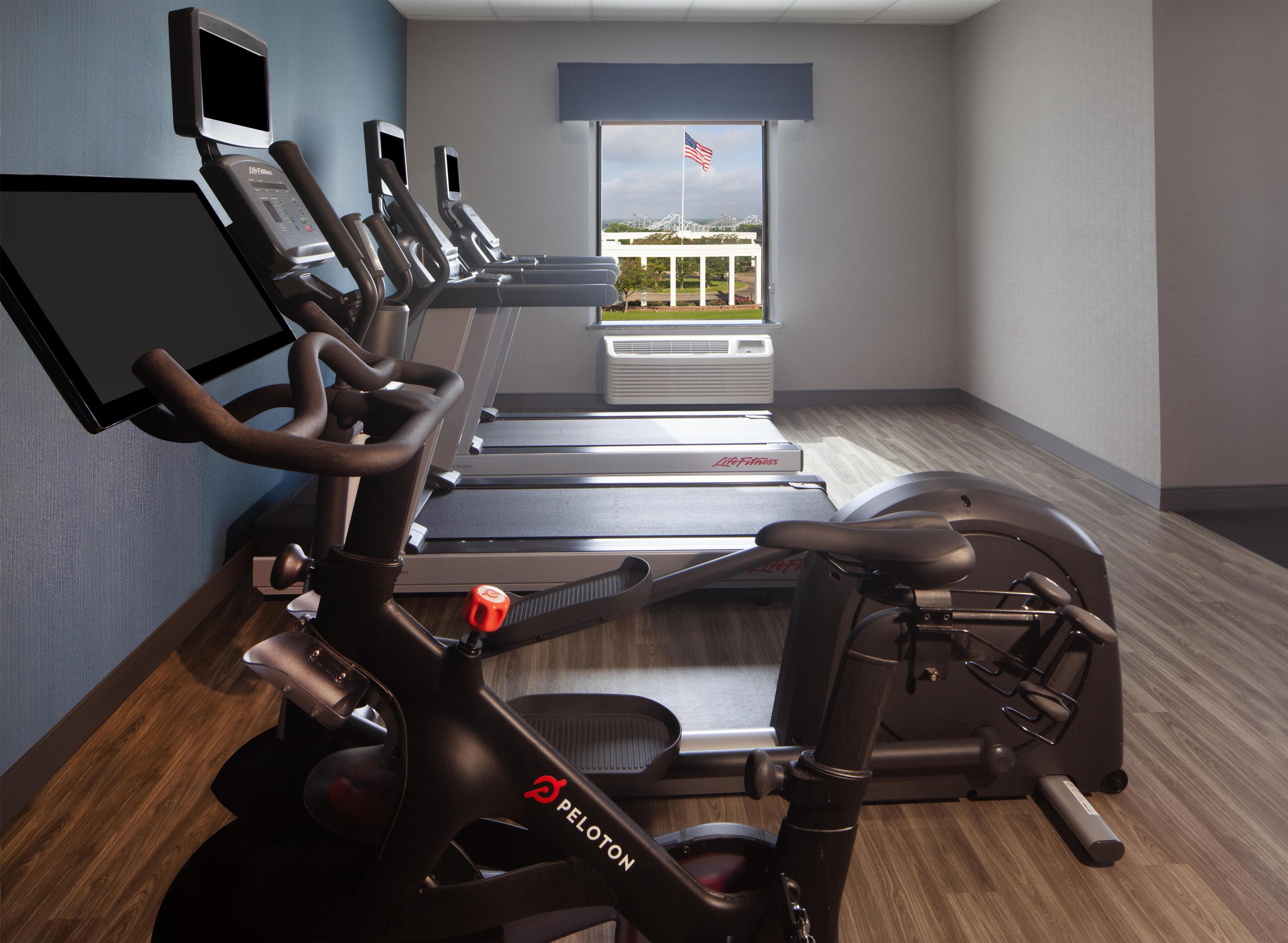Une salle de sport équipée de tapis de course, d’un vélo elliptique et d’un vélo d’appartement, avec une grande fenêtre donnant sur un drapeau américain.