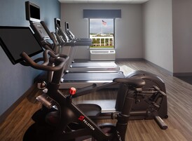 Un gimnasio con cintas de correr, una elíptica y una bicicleta estática, con un amplio ventanal que ofrece vistas de la bandera estadounidense.