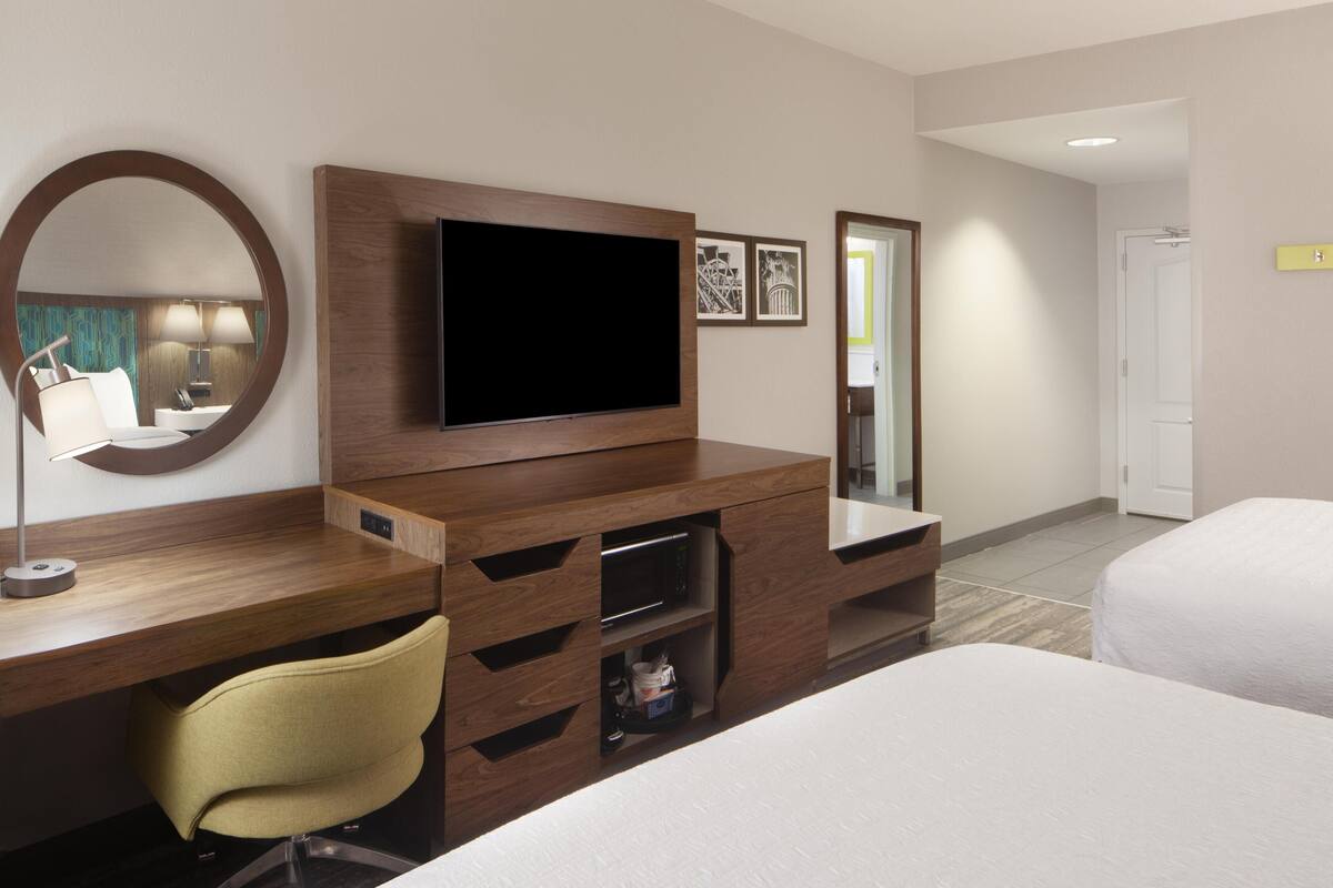 A hotel room with a wooden desk, a television, a mirror, a bed, and a door.