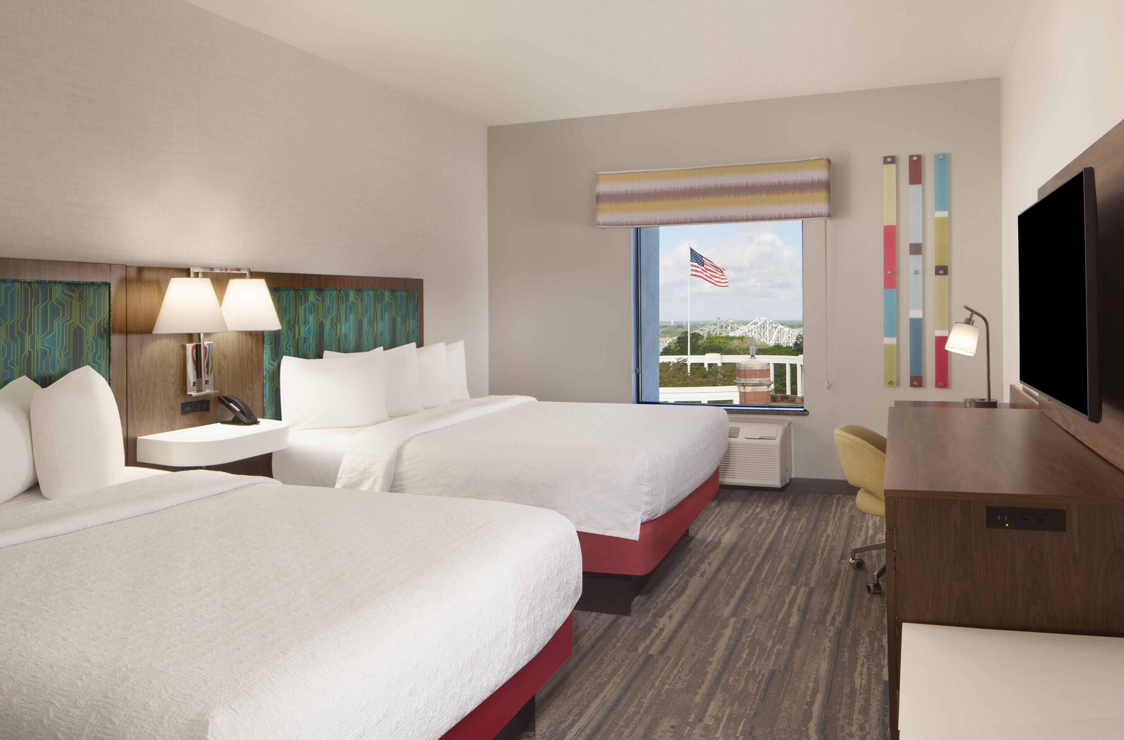 A hotel room with two beds, a desk, a TV, a window with an American flag, and a city view.