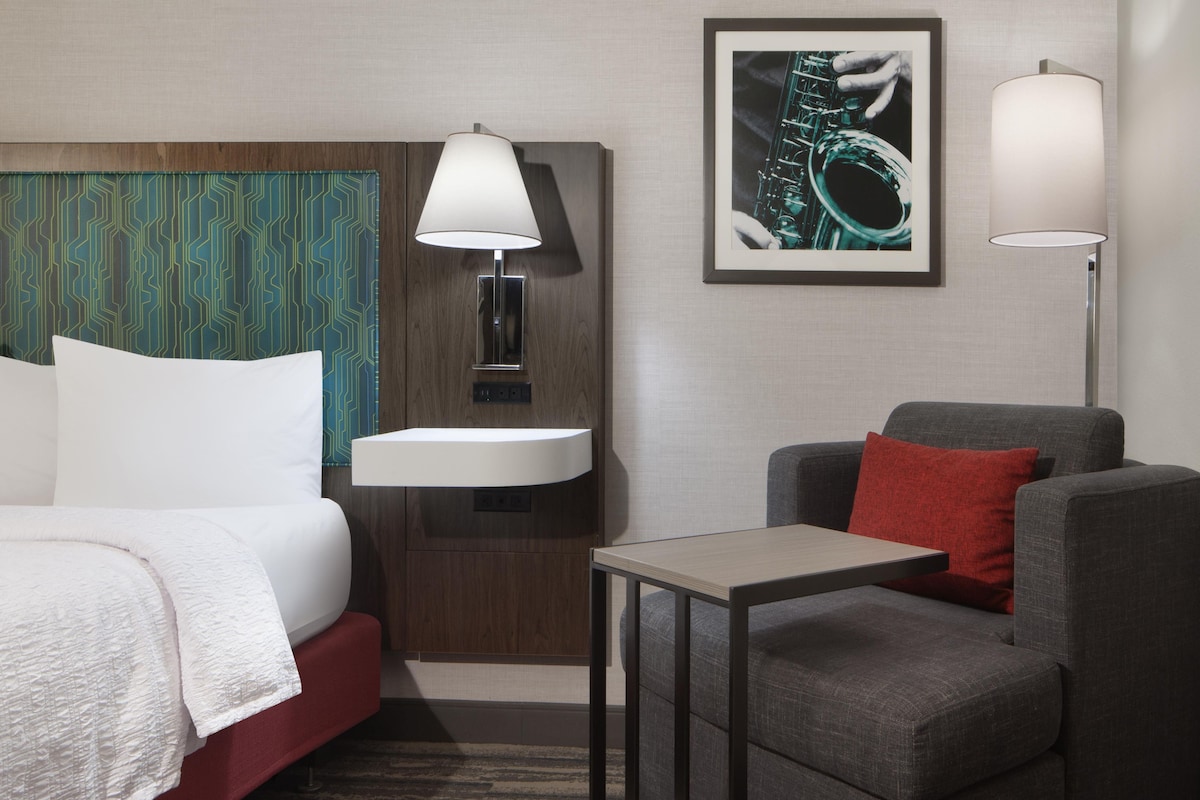 A hotel room with a bed, a bedside table, a chair, a small table, a wall-mounted sink, and a framed picture.