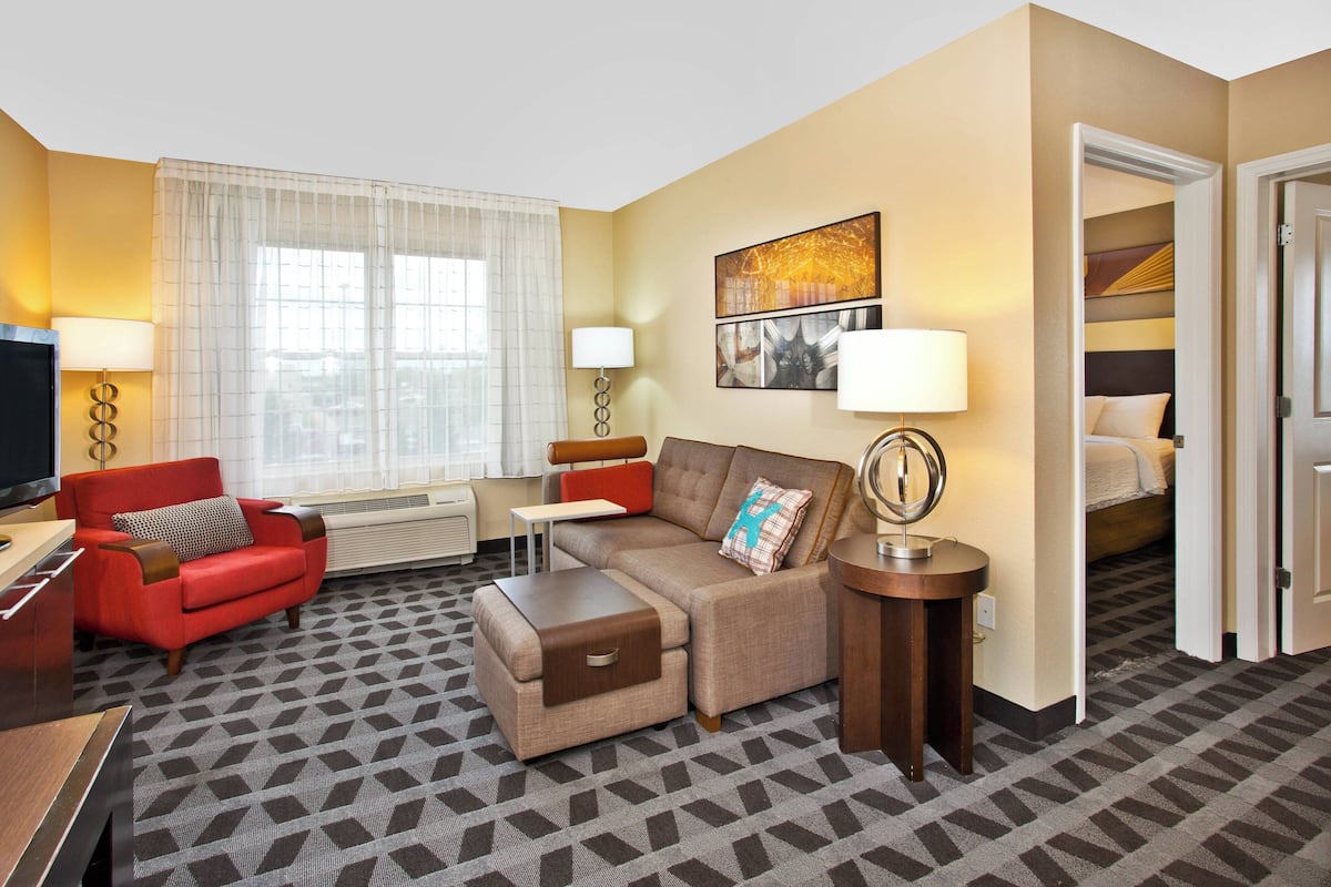 Ein Hotelzimmer mit Sofa, Sessel, Hocker, Fernseher, Schreibtisch und Lampe.
