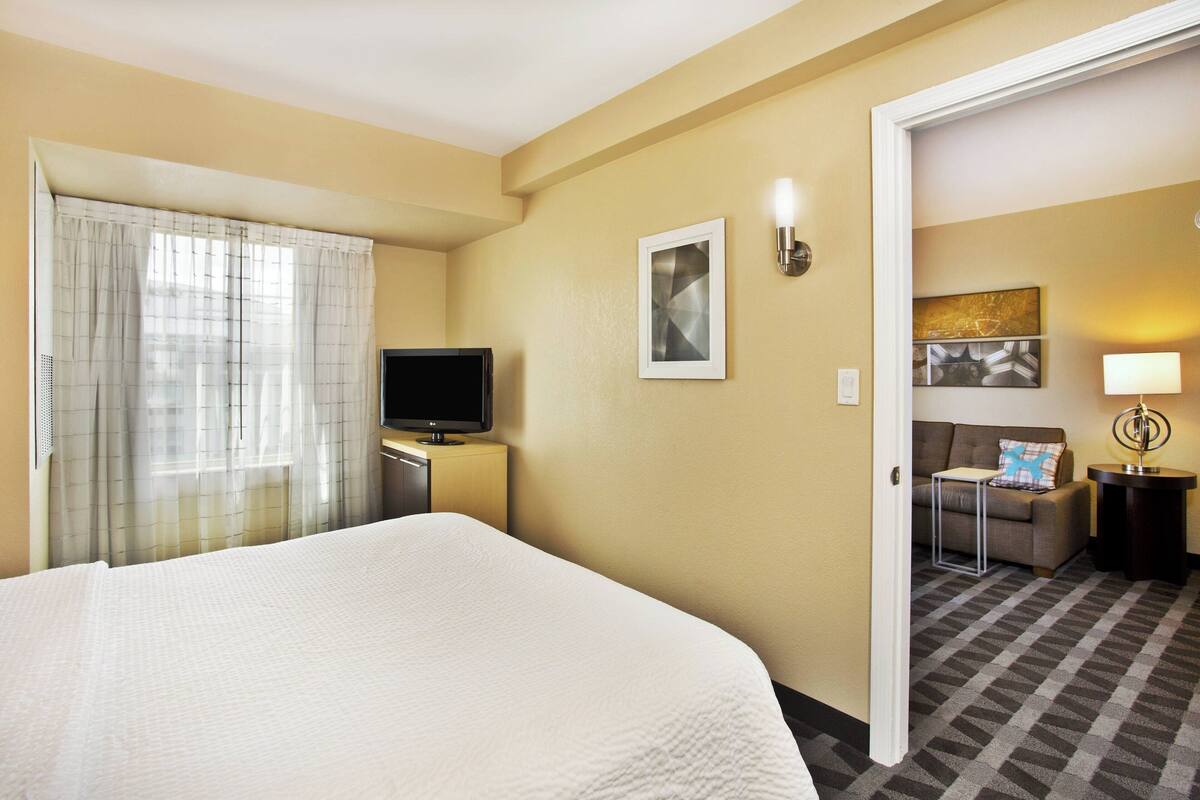Ein Hotelzimmer mit Bett, Fernseher auf einem Schrank, einem Bild an der Wand und einer Couch im angrenzenden Wohnbereich.