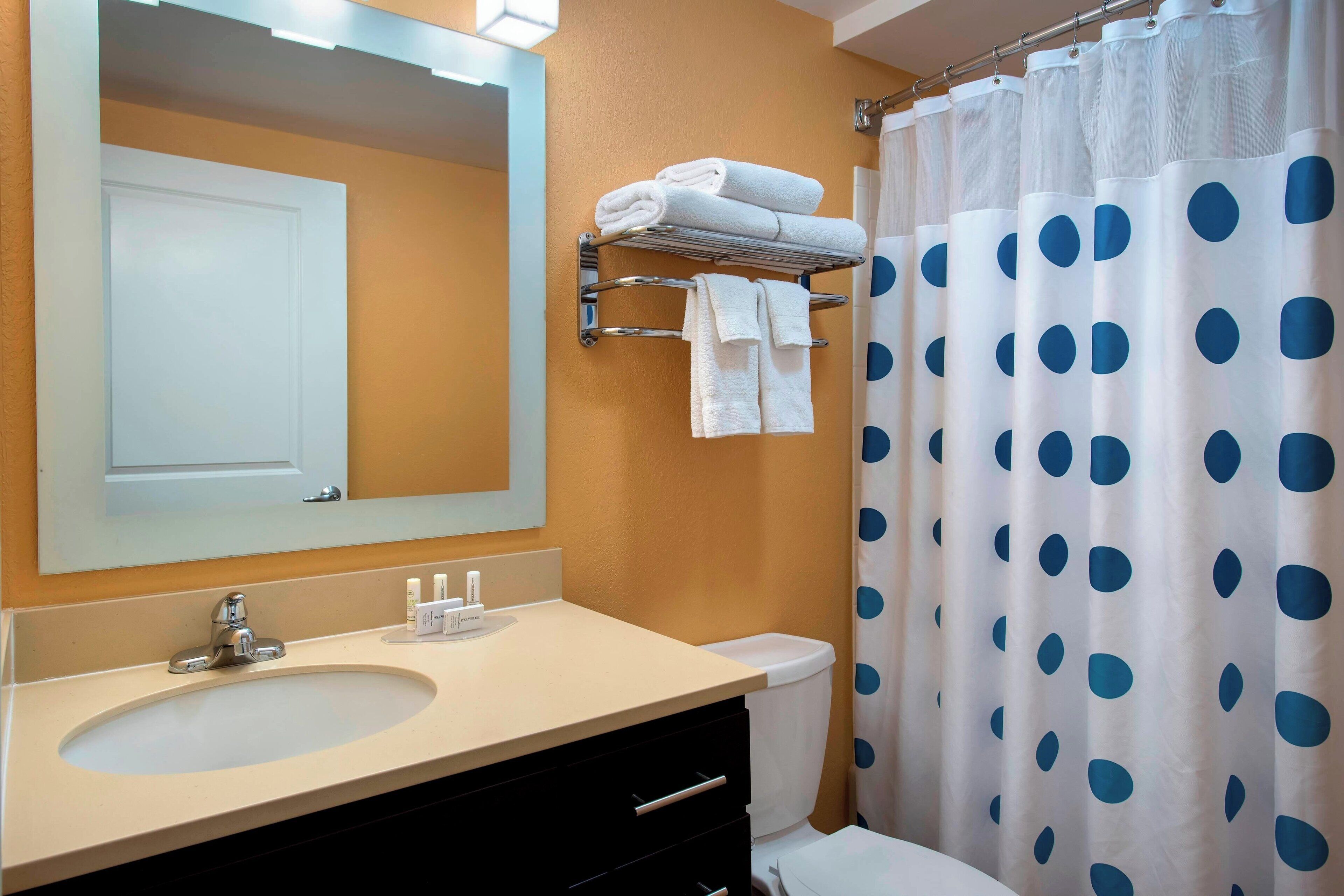 Un baño con un lavabo color crema, un espejo, un perchero con toallas dobladas, un inodoro y una cortina de ducha con lunares.