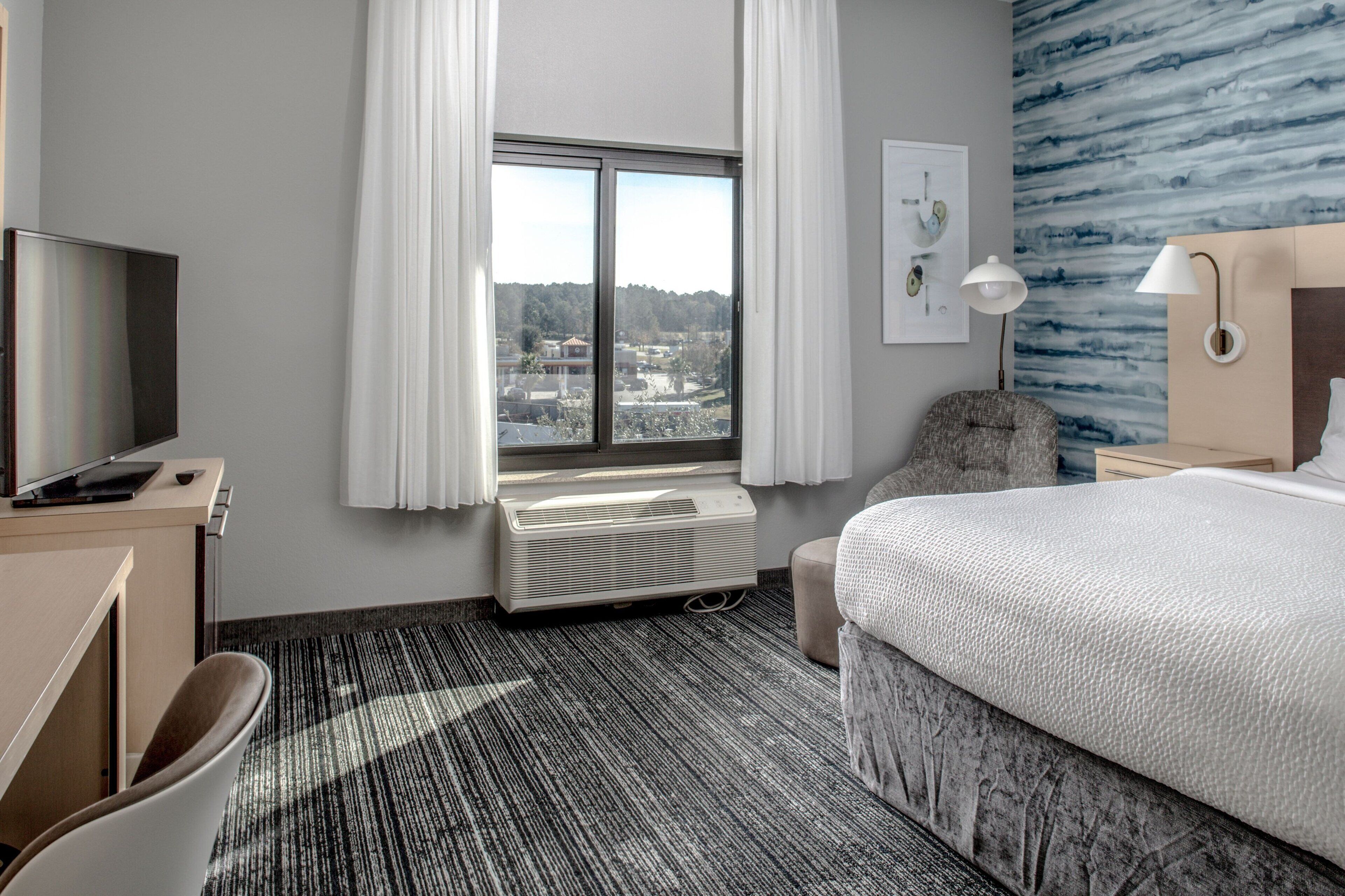 Quarto de hotel com cama, escrivaninha com televisão, uma cadeira, janela com cortinas e parede com padrão azul.
