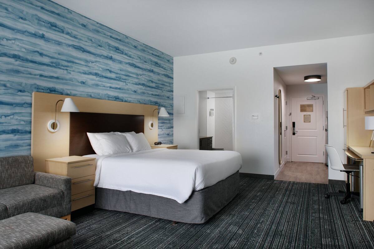 A hotel room with a large bed, a desk, a chair, a sofa, and a bathroom visible through an open door.