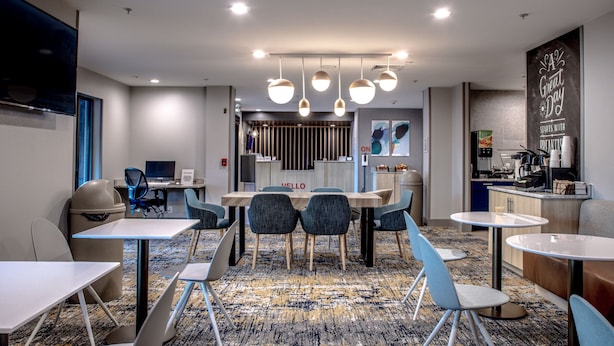 Un hall d’hôtel moderne avec un espace de restauration central, un comptoir d’accueil et une cuisine visible au fond.