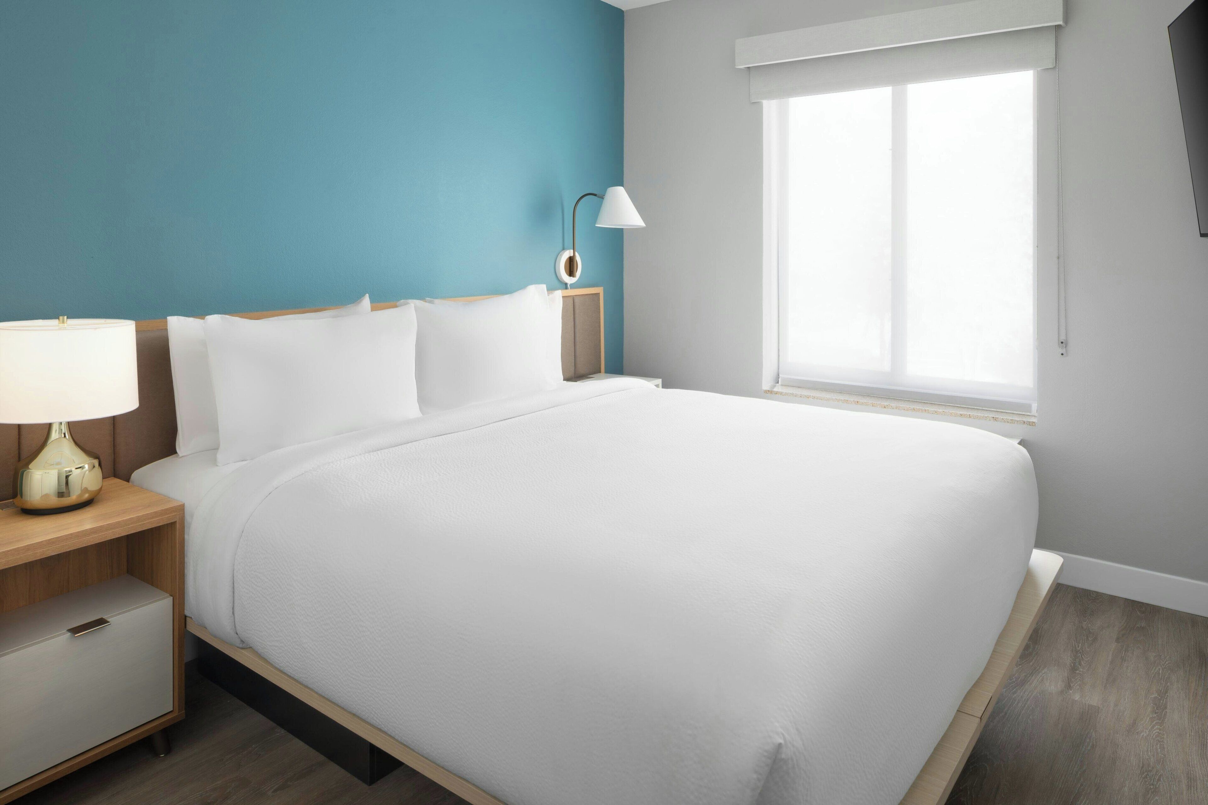 Una cama bien hecha en una habitación con una pared azul decorativa, una lámpara de noche y una ventana con persianas.