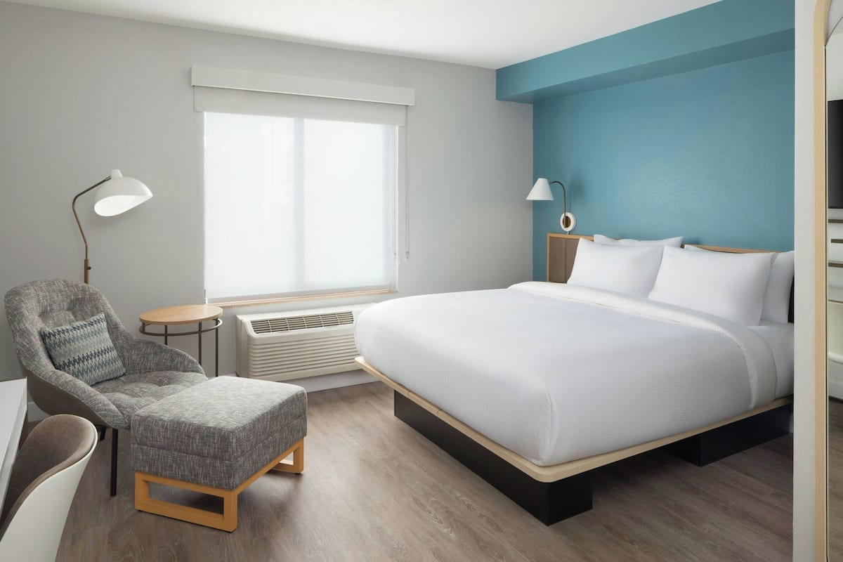 A modern hotel room with a large bed, a grey armchair, a small round table, and a desk.