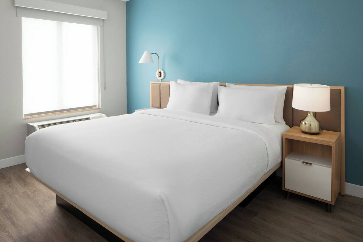 A neatly made bed in a room with a blue wall, a bedside table with a lamp, and a window with blinds.