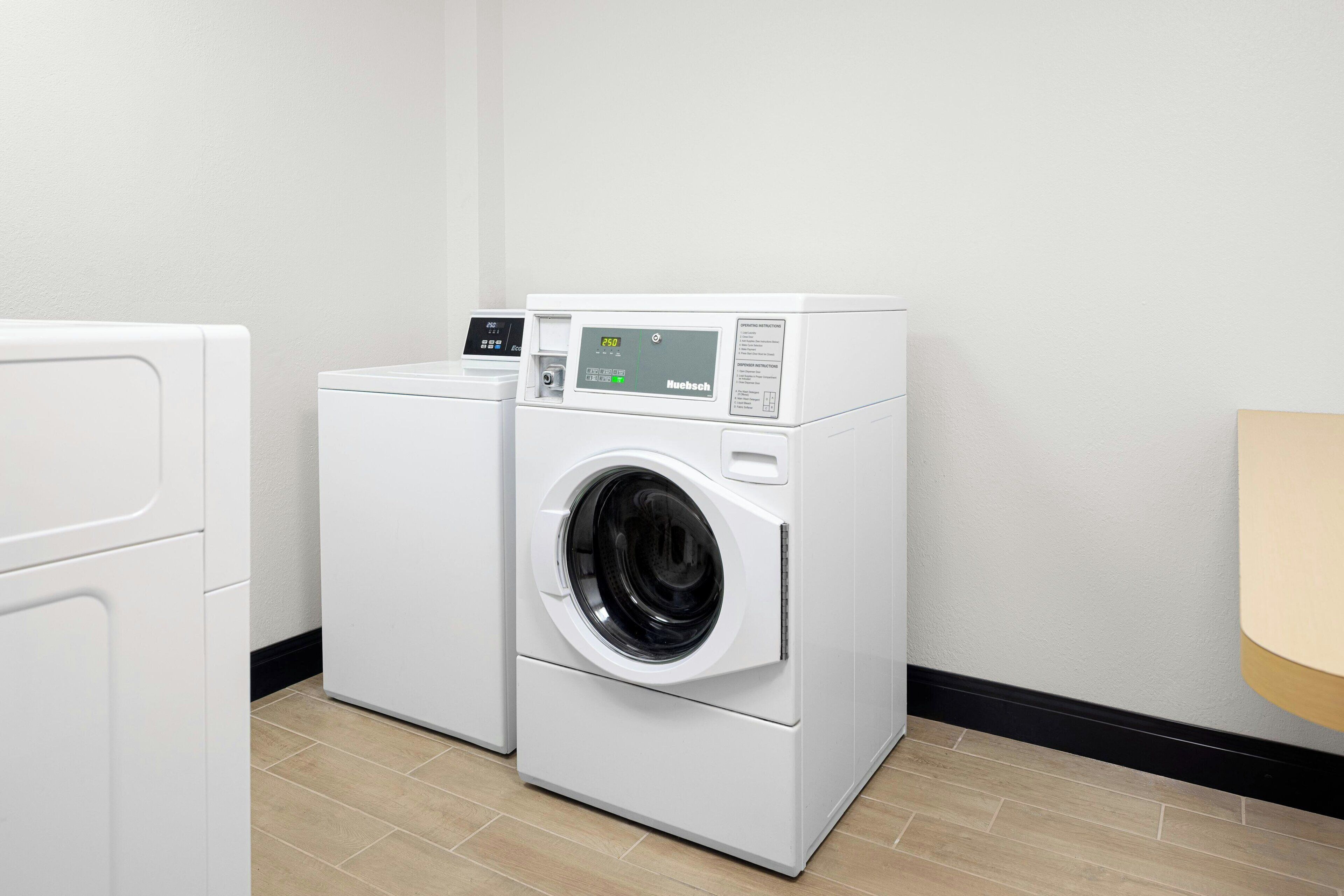 A laundry room with a front-loading washing machine and dryer, a control panel, and a wooden floor.