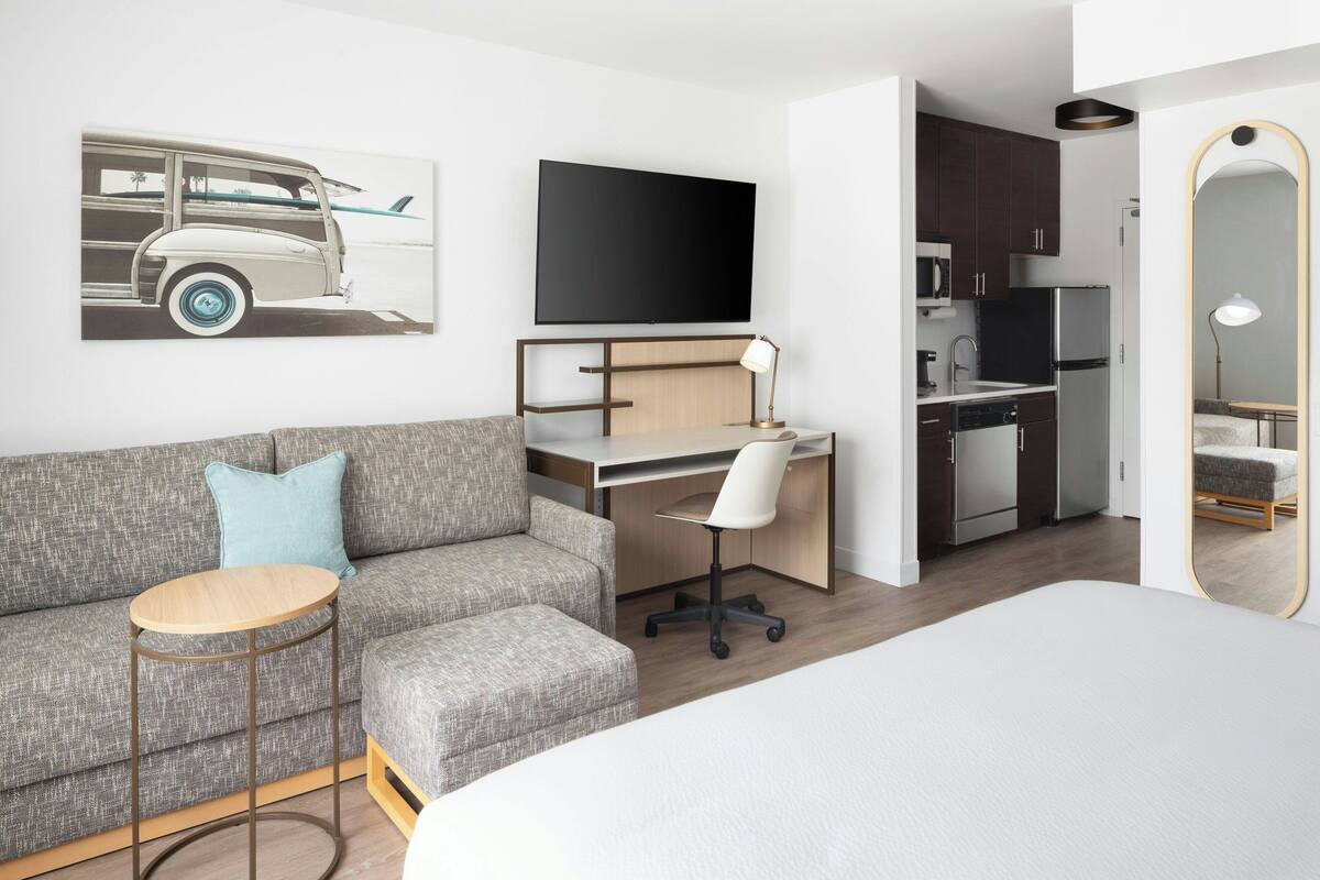 A modern hotel room with a grey sofa, a small round table, a desk with a chair, a flat-screen TV, a kitchen area, and a large mirror.