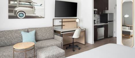 Una habitación de hotel moderna con un sofá gris, una mesita redonda pequeña, un escritorio con una silla, un televisor de pantalla plana, una zona de cocina y un espejo grande.