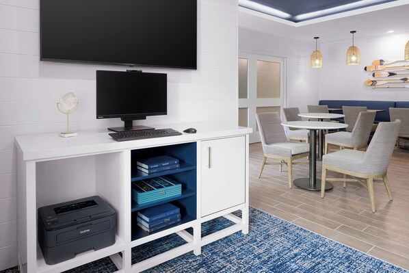 A modern hotel room with a flat-screen TV, a desk with a computer, a printer, and a bookshelf. There are multiple seating areas with tables and chairs.