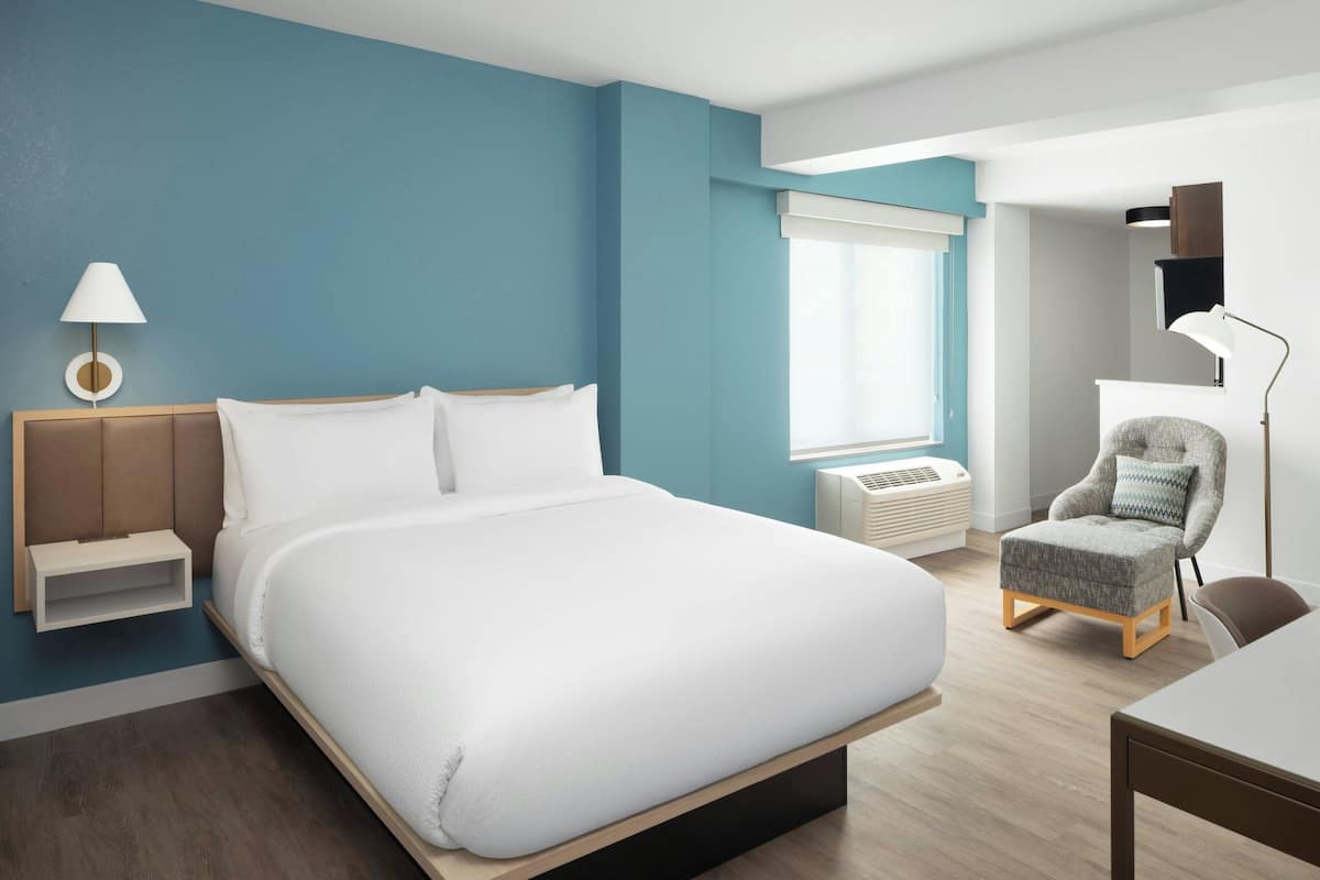 A modern hotel room with a large bed, a bedside table with a lamp, a chair, and a desk.