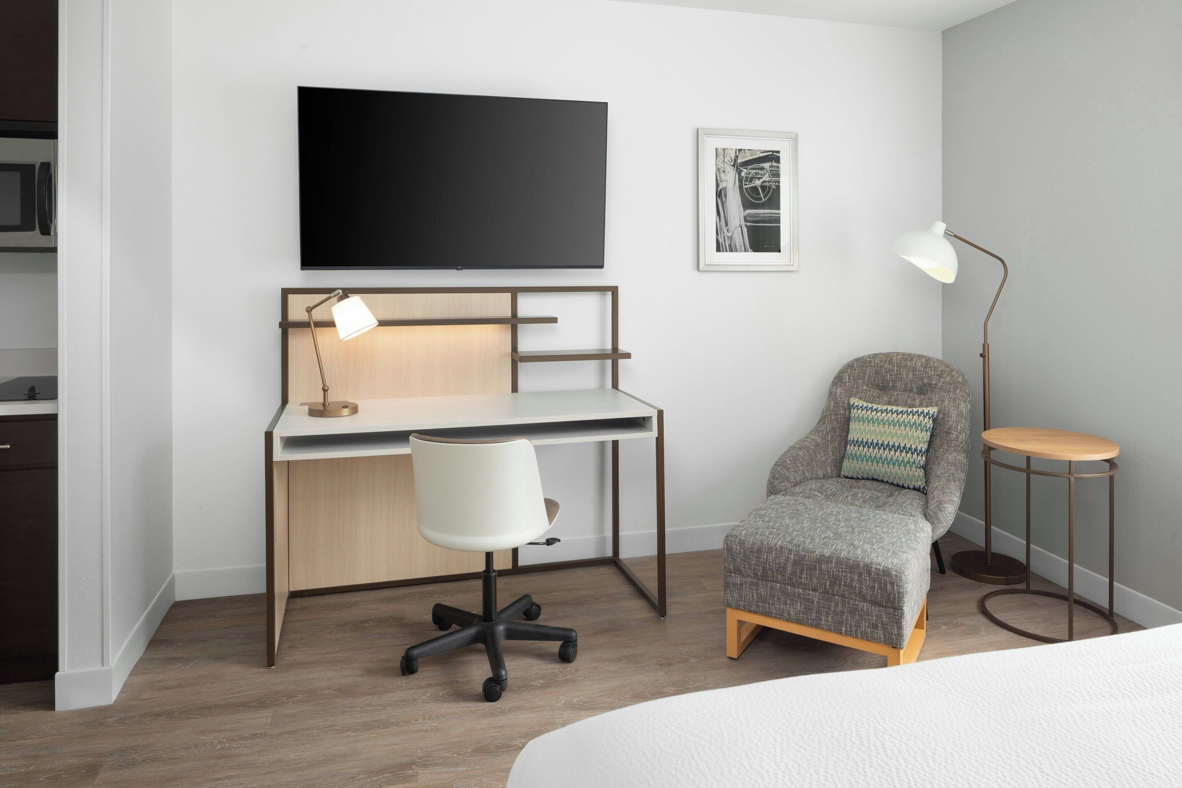 A modern hotel room with a wooden desk, a white office chair, a grey armchair with a patterned cushion, a round side table, a floor lamp, a flat-screen TV mounted on the wall, and a framed picture above the desk.