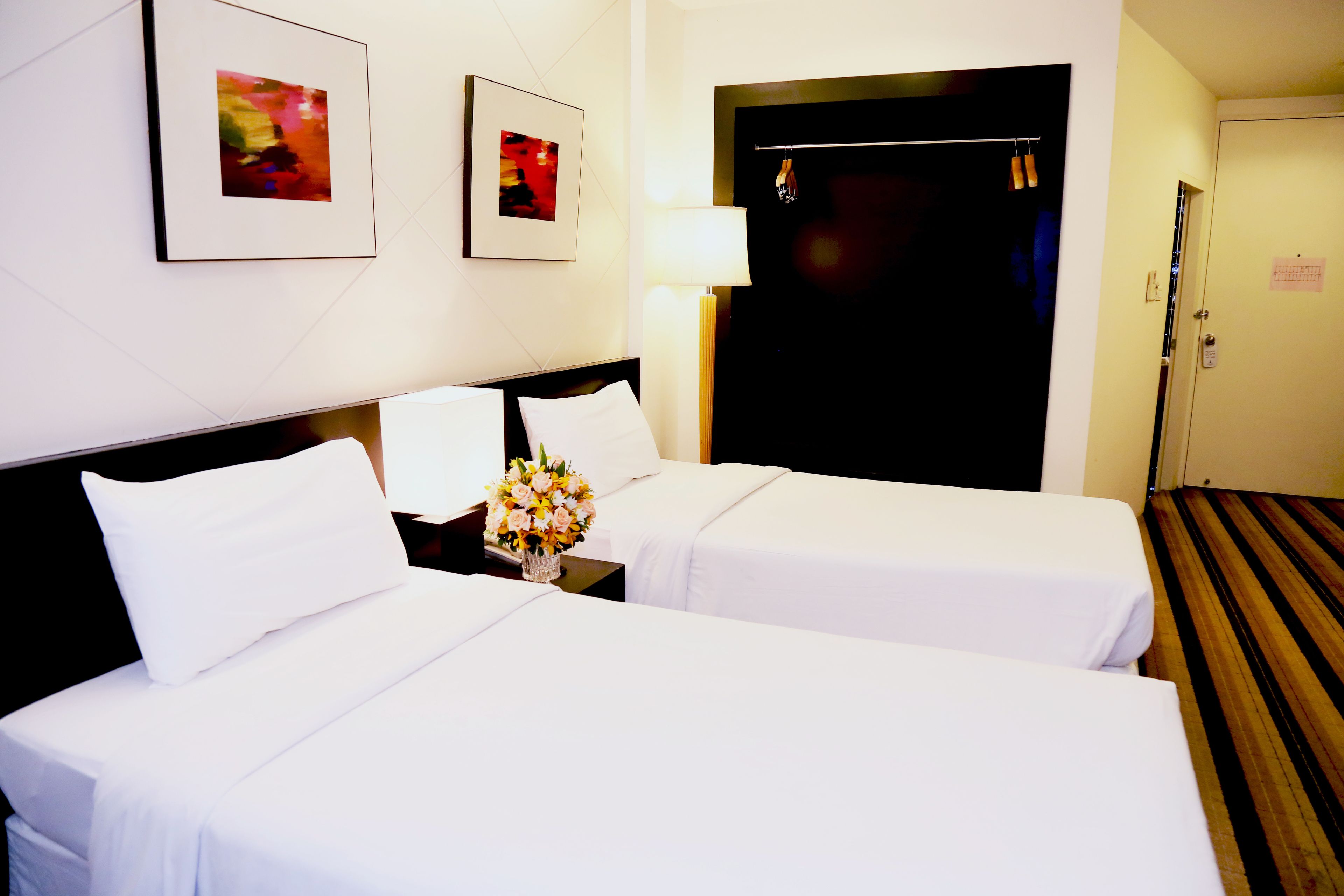 A hotel room with two beds, a nightstand with a lamp and a vase of flowers, and a door with a coat rack.