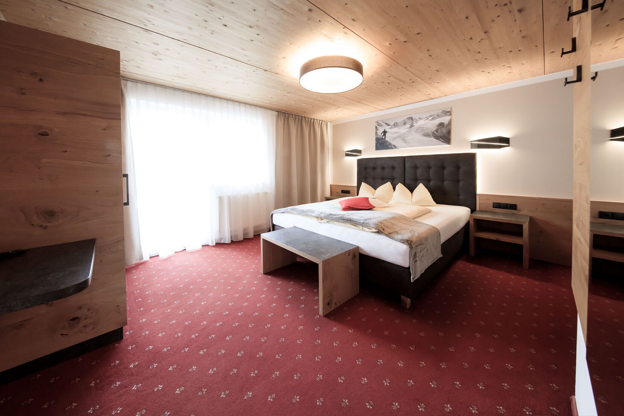 A hotel room with a wooden ceiling, a large bed with a headboard, a bench at the foot of the bed, a nightstand, and a wall-mounted light fixture.