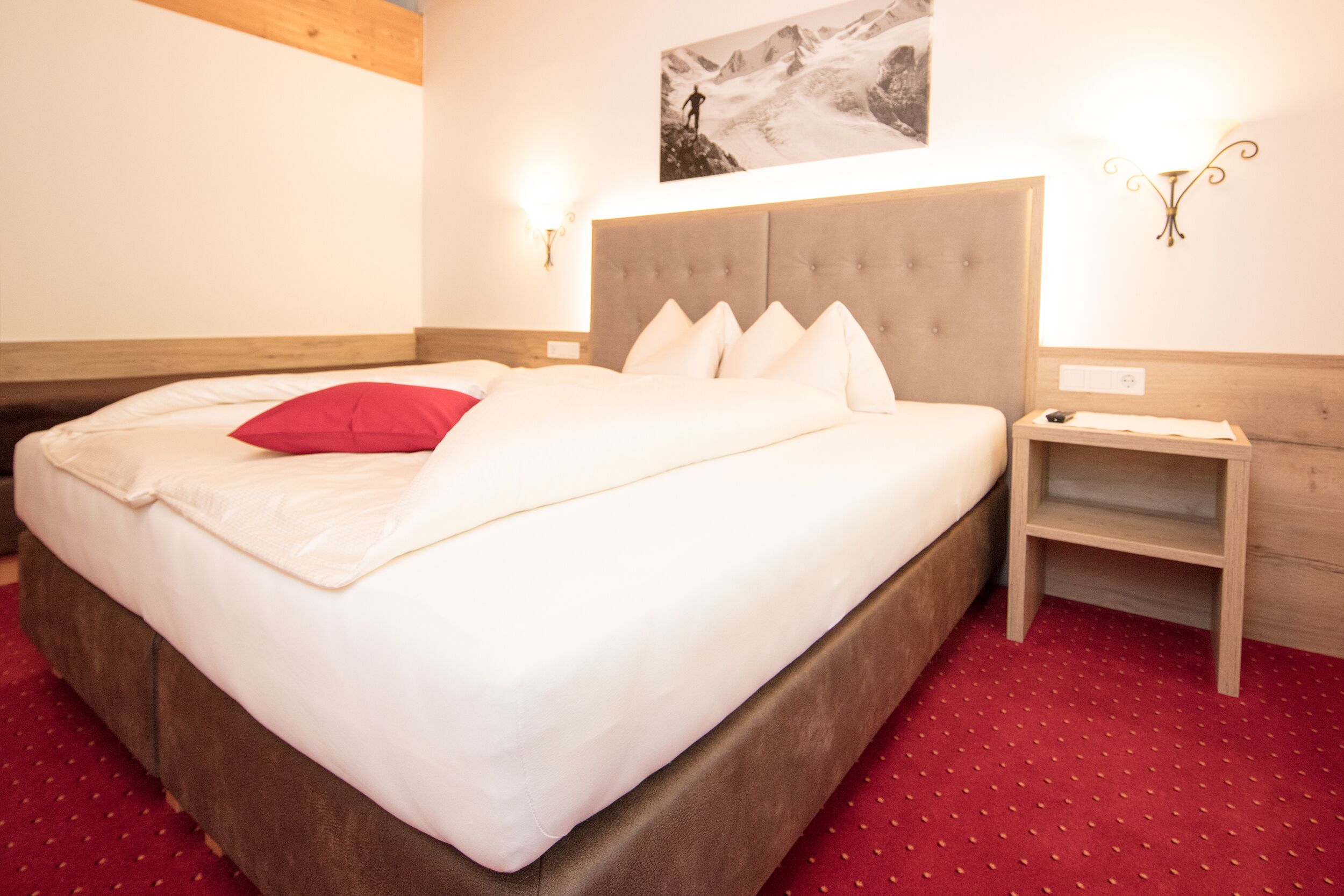 Een hotelkamer met een groot bed, een houten hoofdeinde, een rood tapijt en een nachtkastje met een lamp.