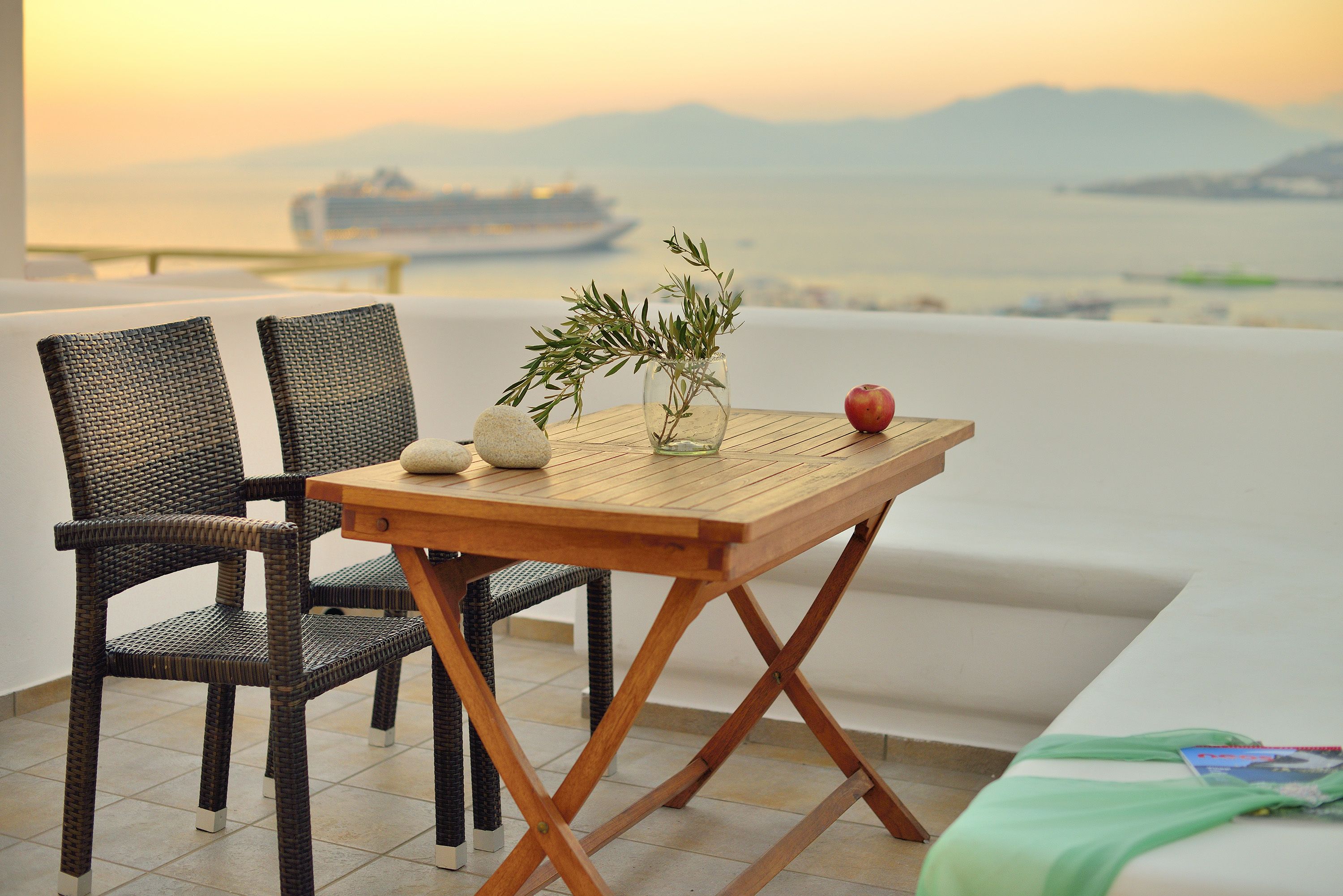 Ein Holztisch mit einer Vase mit Grünpflanzen und einem Apfel, zwei Korbstühle und Blick auf das Meer mit einem Schiff in der Ferne.