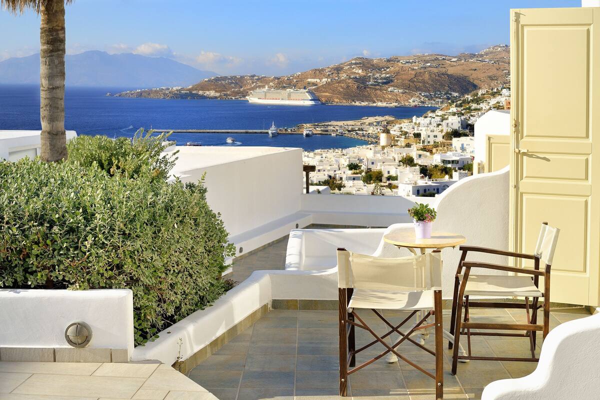A terrace with a table and chairs, a palm tree, and a view of the sea and a cruise ship.