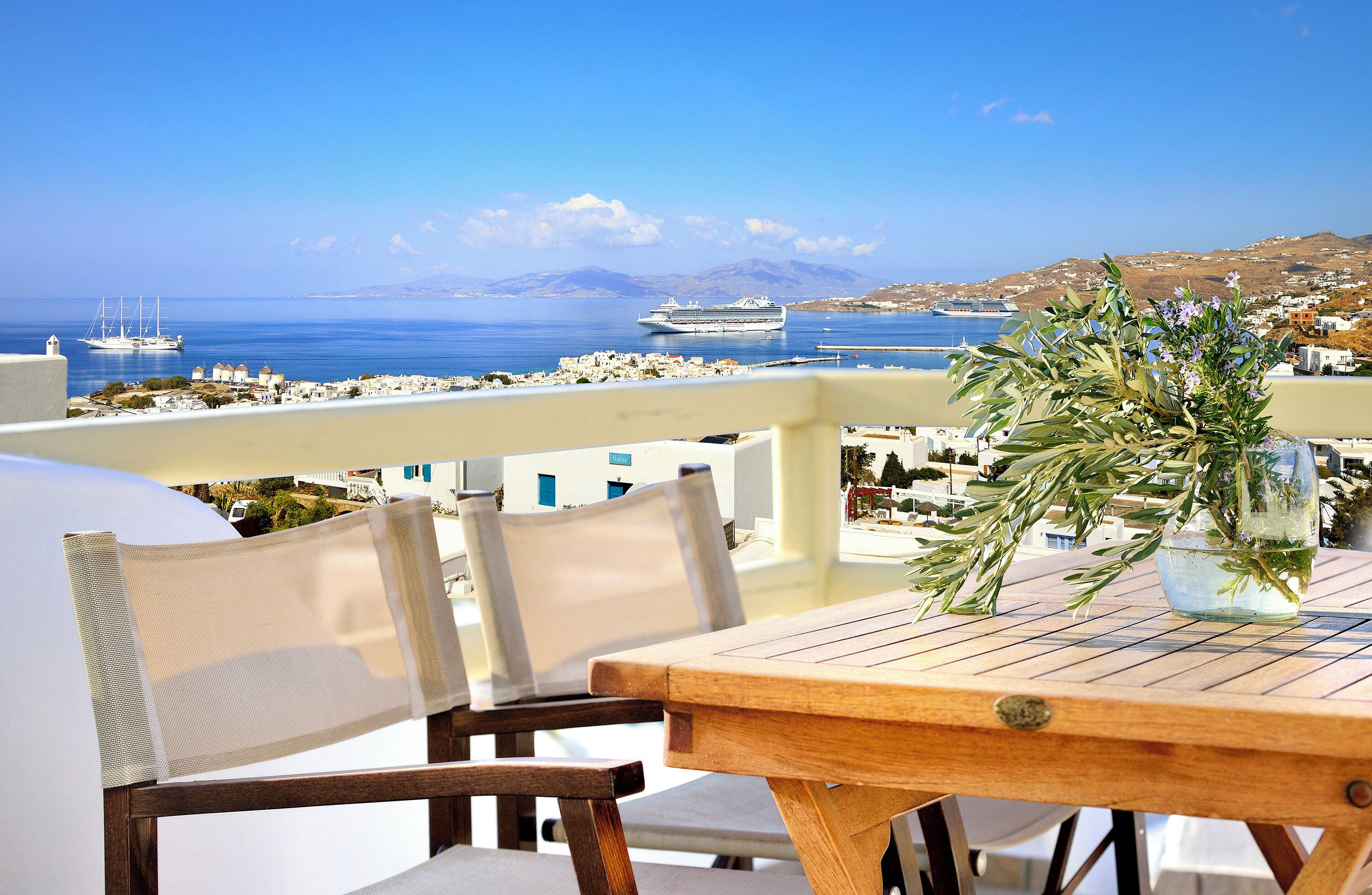 A wooden table with a vase of greenery on a balcony overlooking a coastal town with buildings and boats.