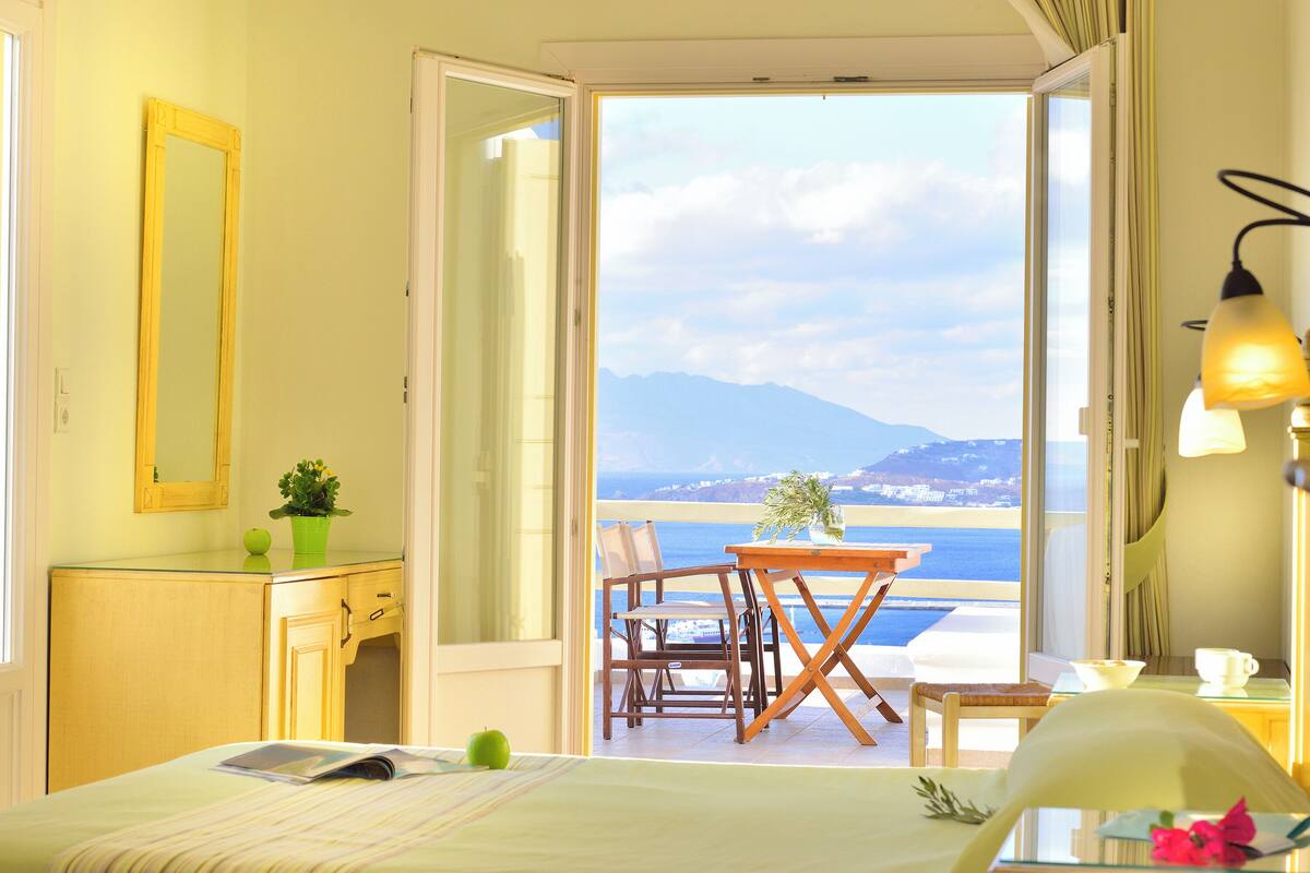 A room with a bed, a desk, a chair, a table, and a balcony with a view of the sea and mountains.