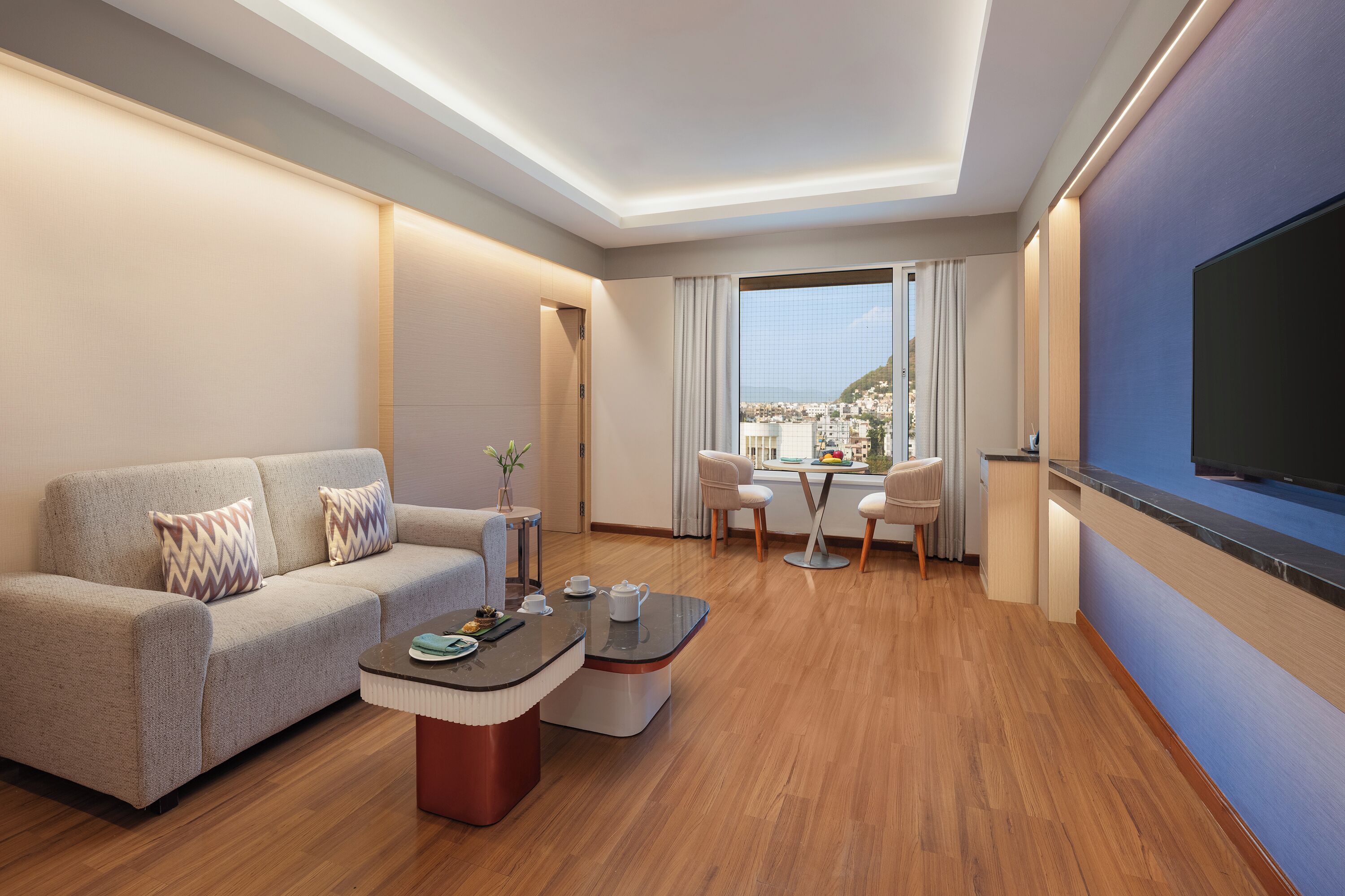 A modern hotel room with a sofa, coffee table, and a TV. There is a balcony with a view of the city.