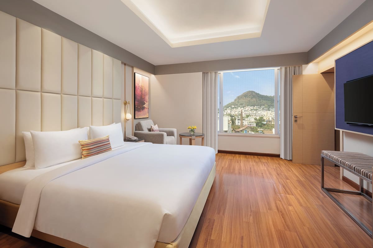 Een hotelkamer met een groot bed, een televisie, een klein tafeltje met bloemen en uitzicht op een stadsbeeld.