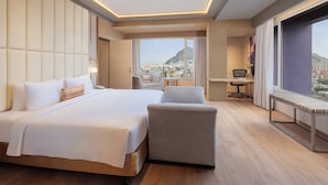 Ein Hotelzimmer mit einem großen Bett, einem Schreibtisch, einem Sessel und Blick auf die Stadt.