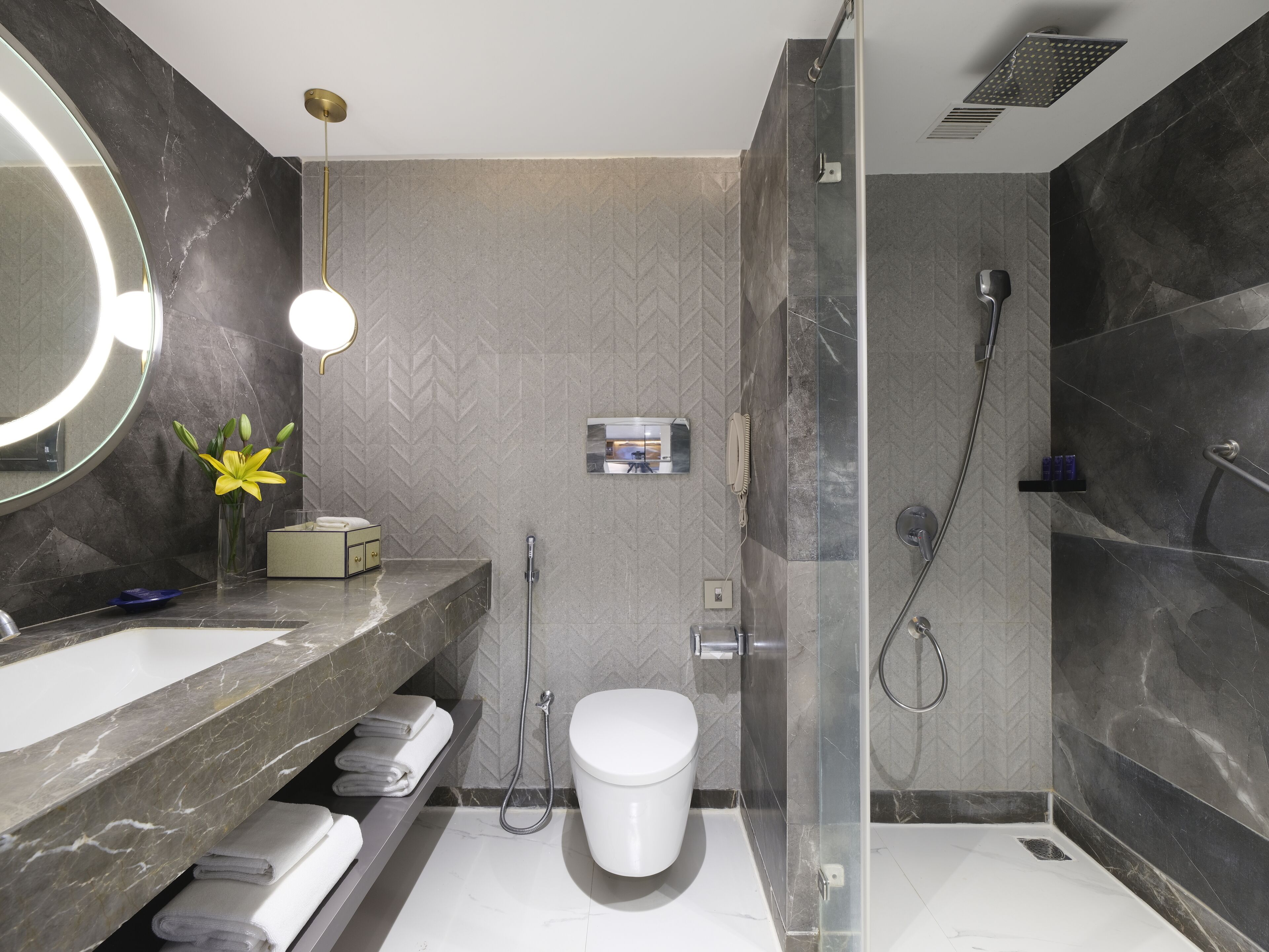 A modern bathroom with a large mirror, a round light fixture, a toilet, a shower, and a vanity with a marble countertop.