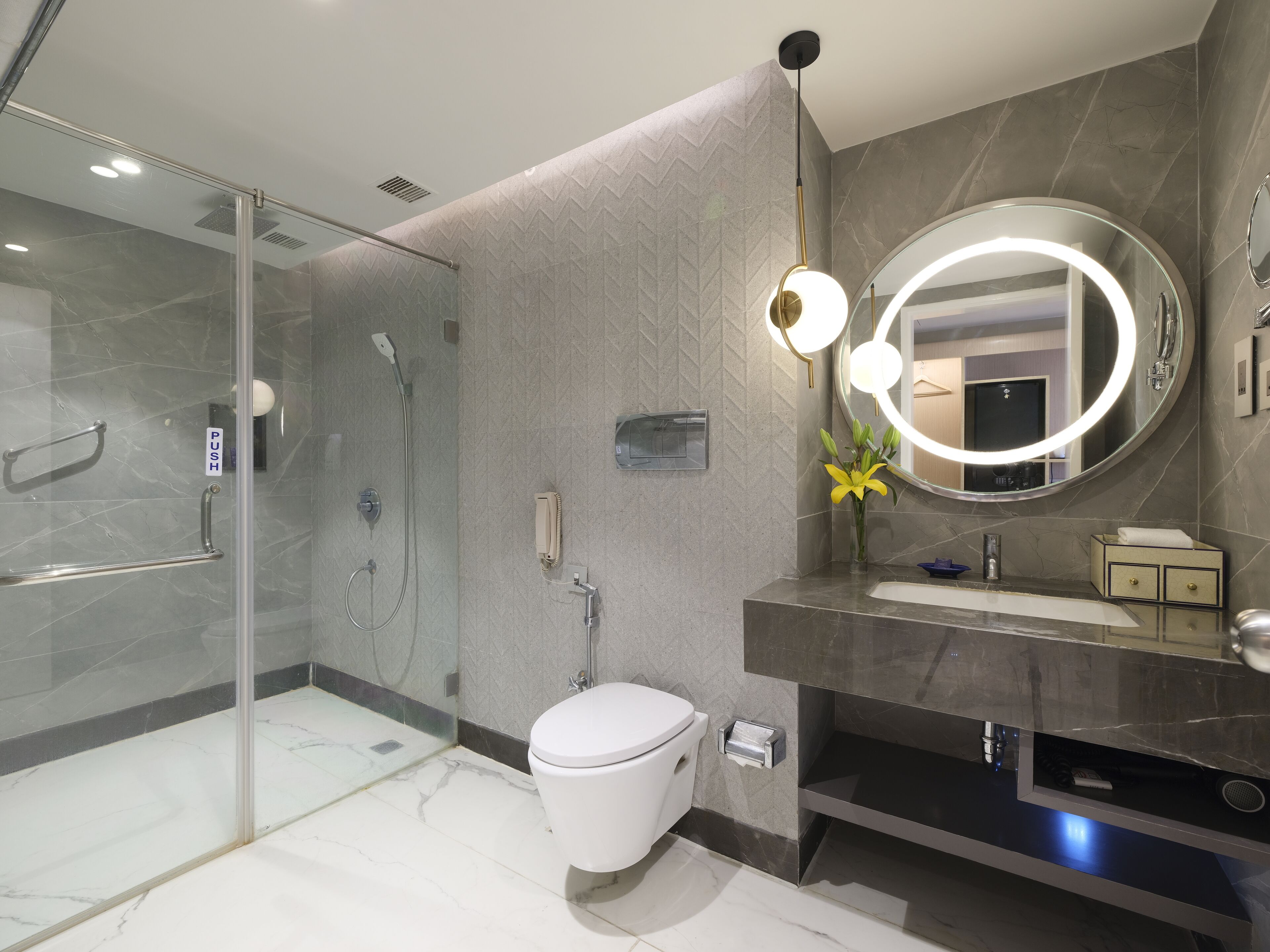 Un baño moderno con cabina de ducha de vidrio, inodoro, lavamanos con tocador y un espejo grande con luz circular.