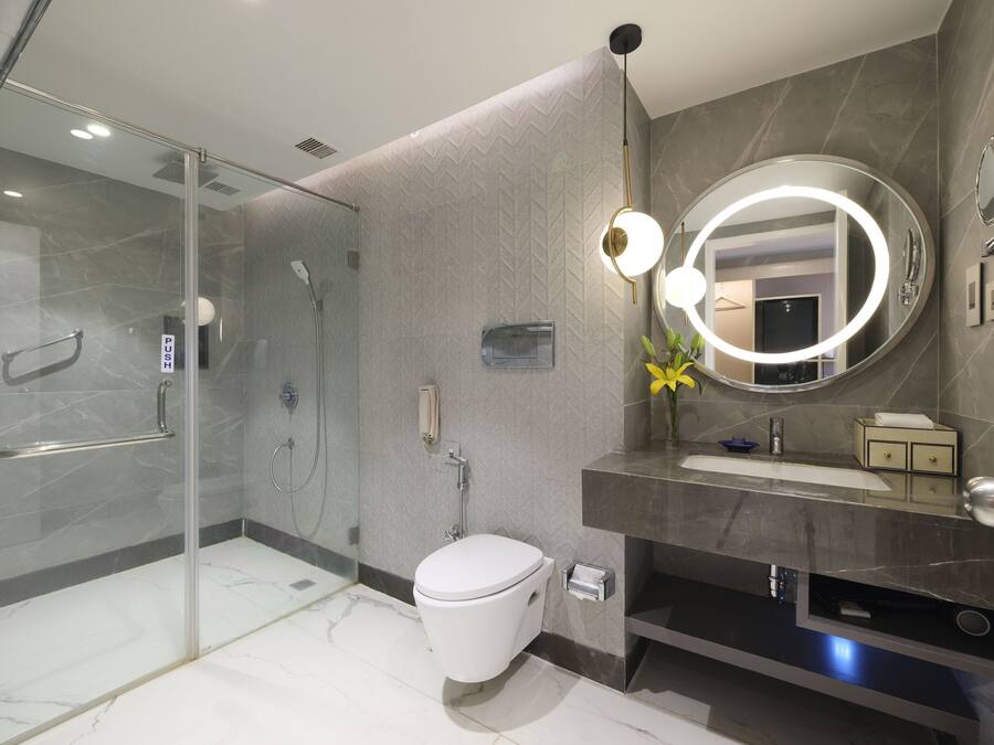 Un baño moderno con cabina de ducha de vidrio, inodoro, lavamanos con tocador y un espejo grande con luz circular.