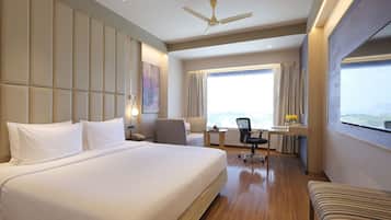 A hotel room with a large bed, a desk with a chair, a ceiling fan, and a large window with a view.