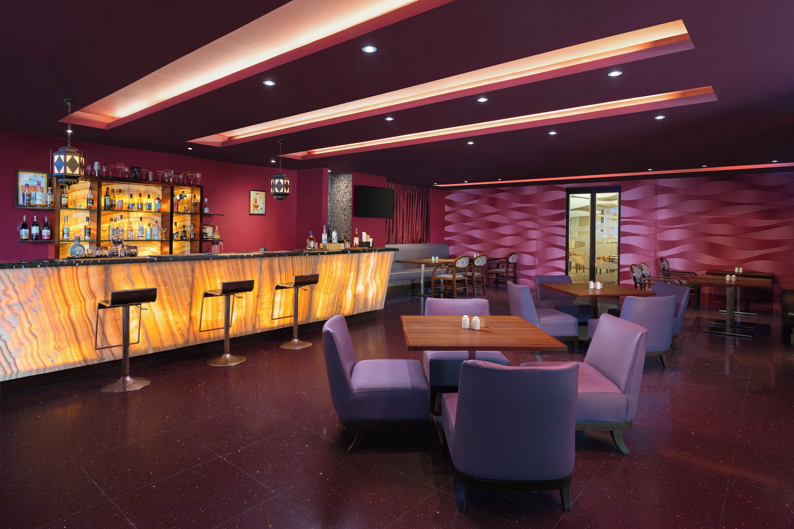 Eine moderne Bar mit Marmorplatte, Barhocker und einer Auswahl an Flaschen. Es gibt Tische und Stühle zum Essen, und die Wände sind mit geometrischen Mustern verziert.
