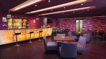 A modern bar area with a marble countertop, bar stools, and a variety of bottles. There are tables and chairs for dining, and the walls are decorated with geometric patterns.
