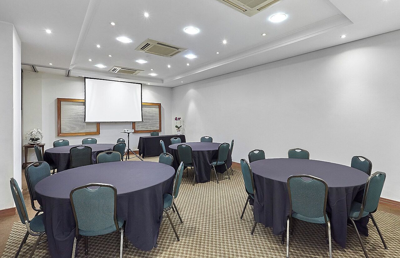 Uma sala de conferências com mesas redondas, cadeiras, uma tela de projetor e piso coberto de carpete.